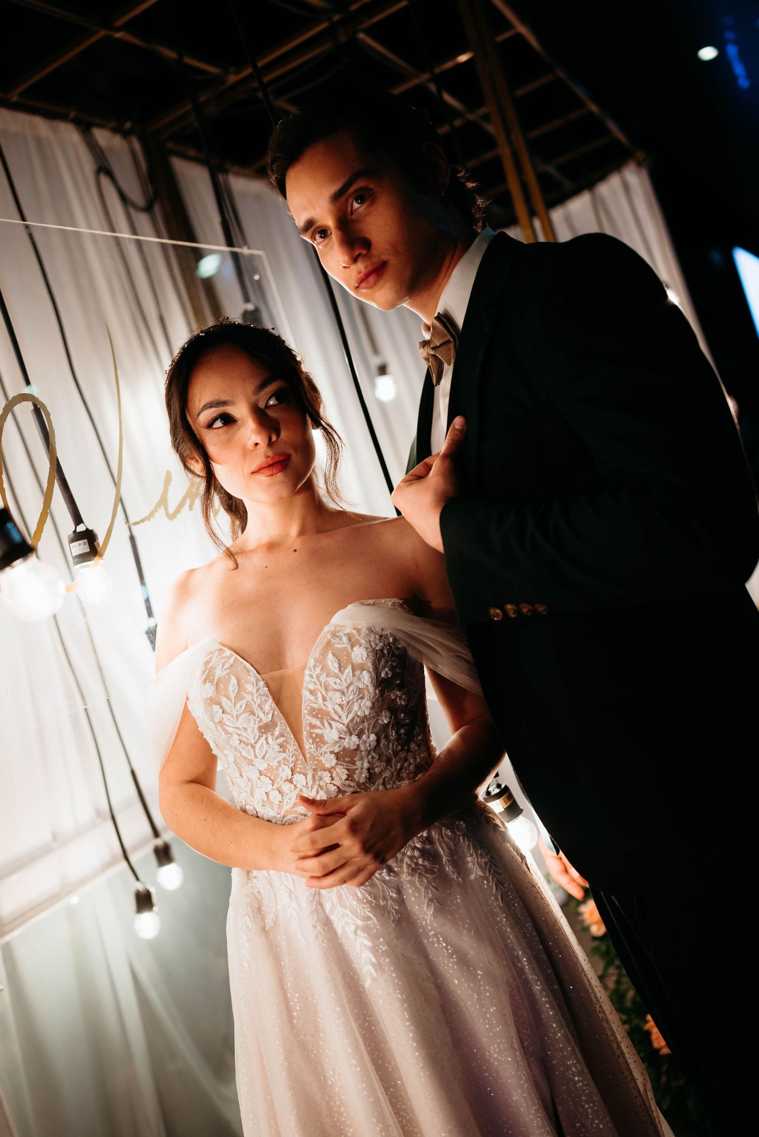 A romantic wedding couple embraces in dramatic low-light photography with hanging lights in the background.