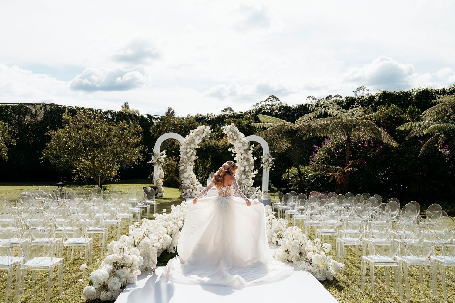 White chairs arranged for an outdoor garden wedding ceremony under an elegant floral arch at a scenic hilltop venue.