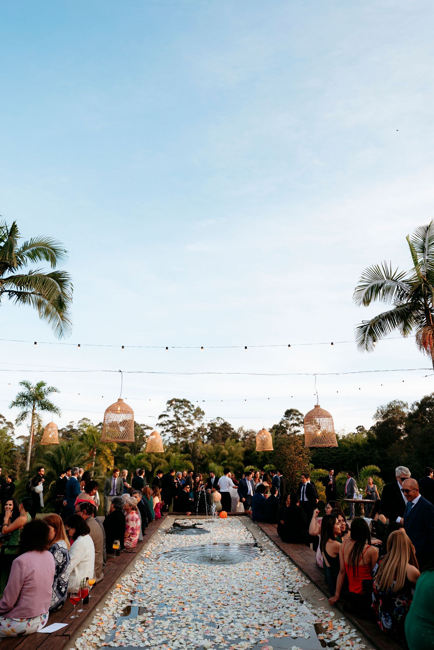 Outdoor wedding reception at dusk with string lights, palm trees, and guests gathered around a floral aisle.