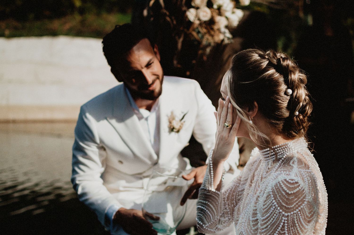 Groom in white suit reads vows to bride in beaded lace wedding dress during outdoor ceremony near water.