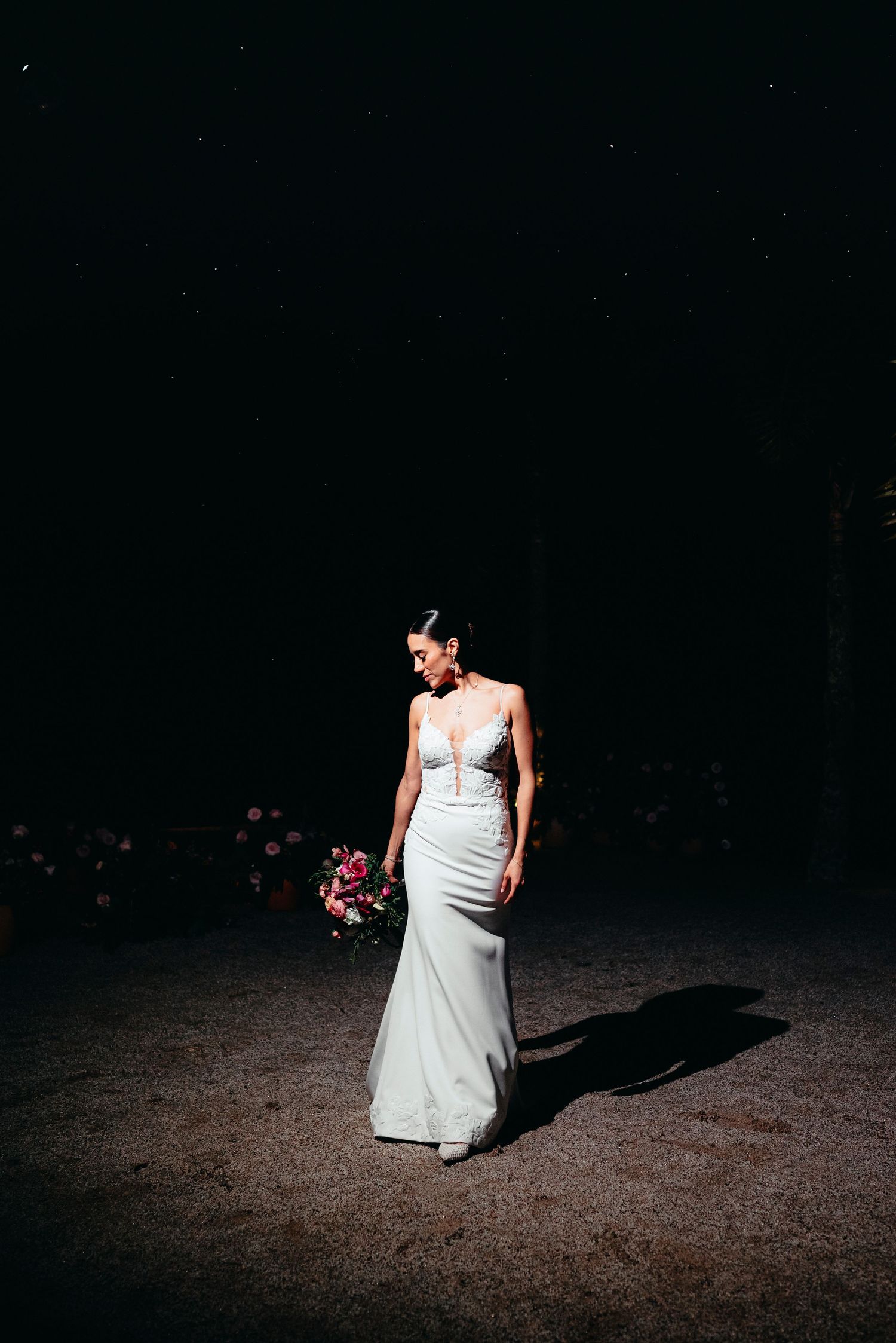 Bride in fitted white lace gown holds colorful bouquet under dramatic spotlight in dark venue.