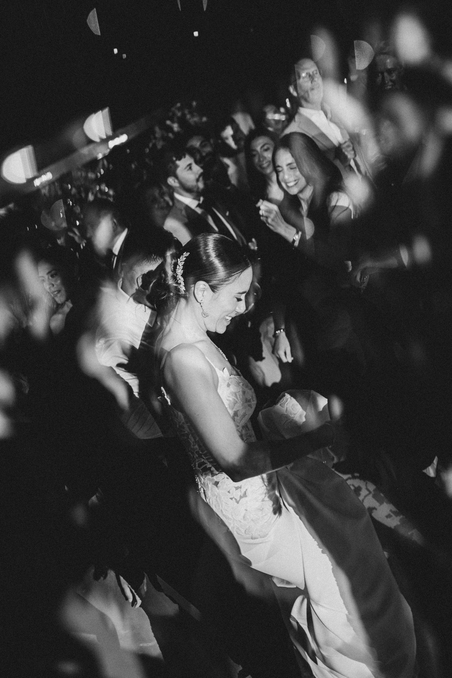 Bride in lace wedding gown dances joyfully surrounded by cheering guests in black and white reception photo.