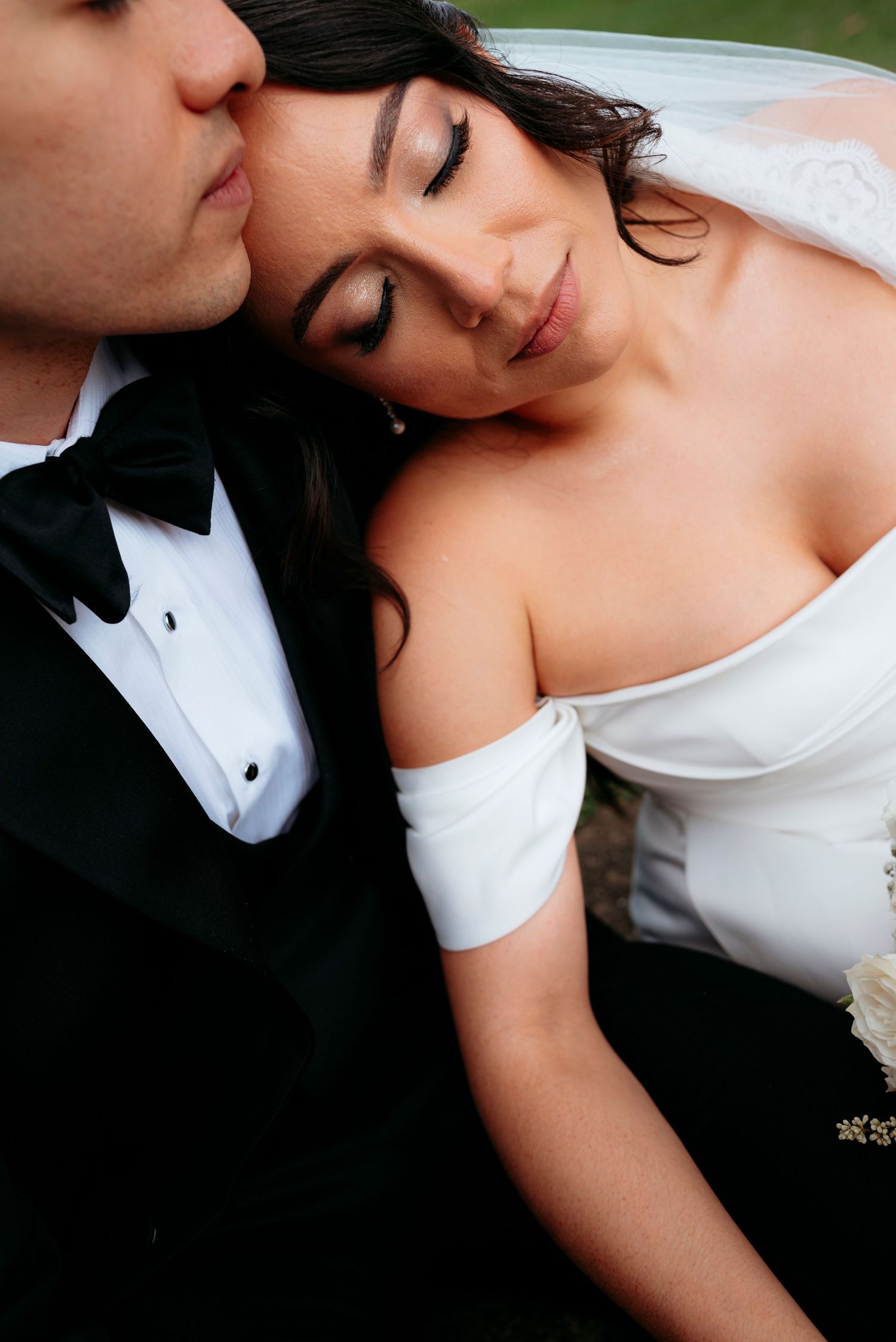 Groom in black tuxedo kisses bride wearing off-shoulder white gown as she rests her head on his chest.