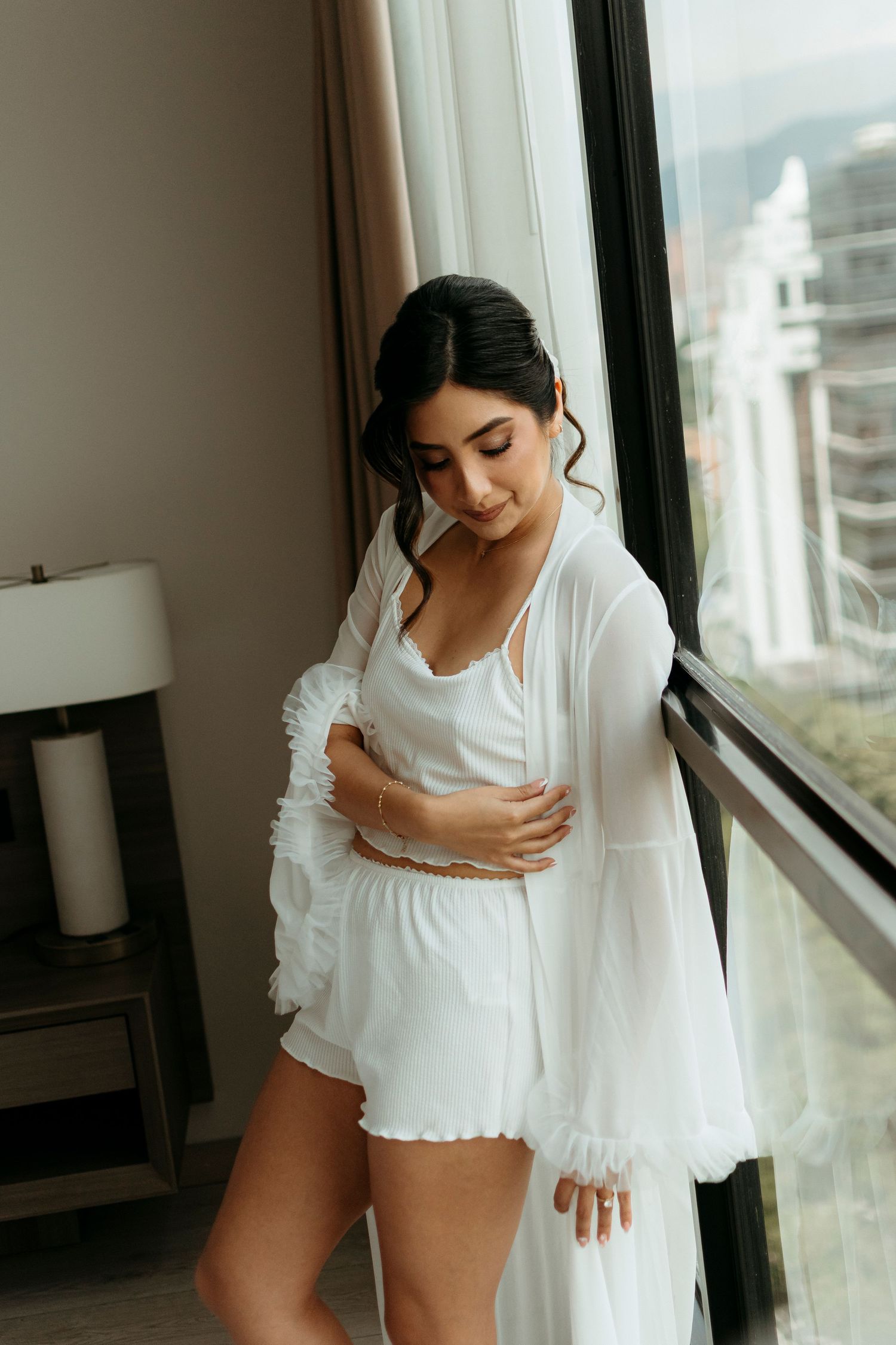 A person in white loungewear stands thoughtfully by a window in a high-rise apartment with city views.