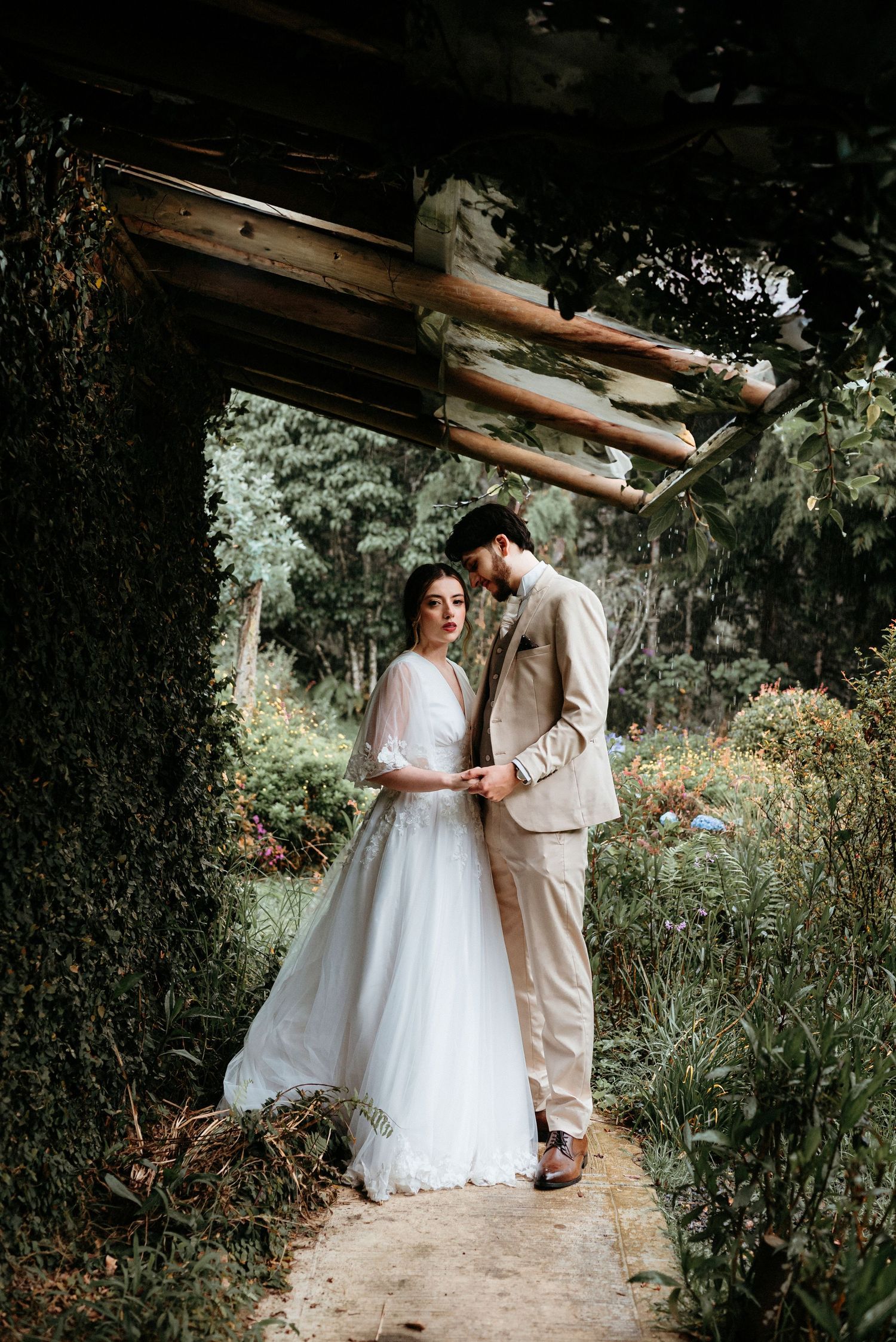 A romantic garden wedding portrait shows a couple embracing under a rustic wooden archway.