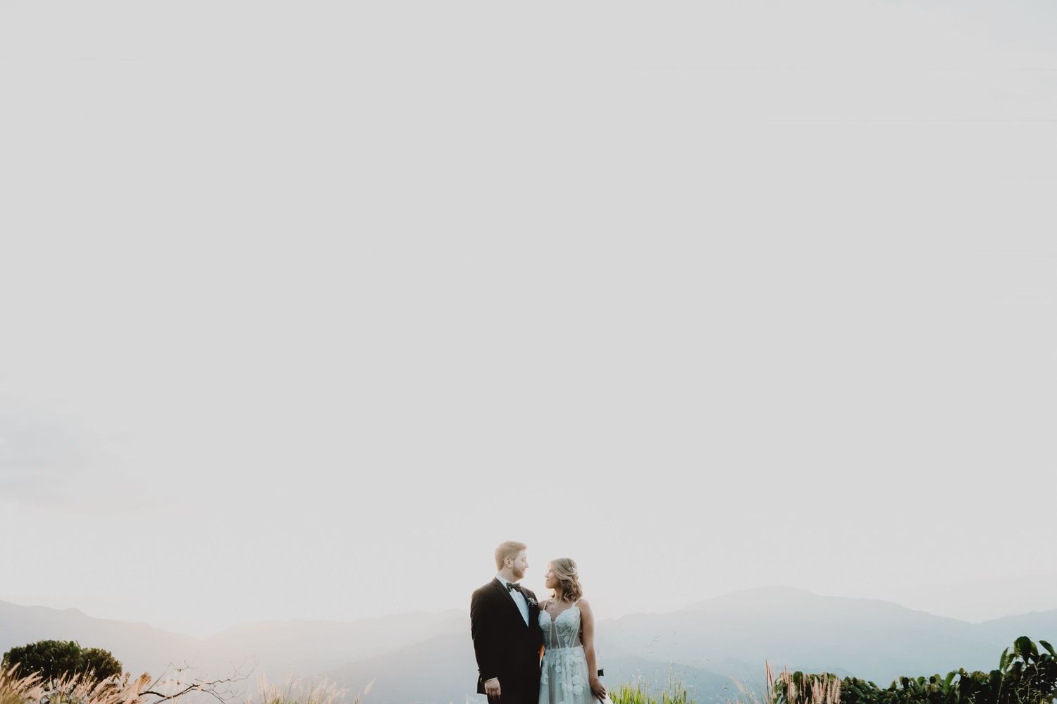 A romantic wedding couple stands silhouetted against a misty mountain backdrop.
