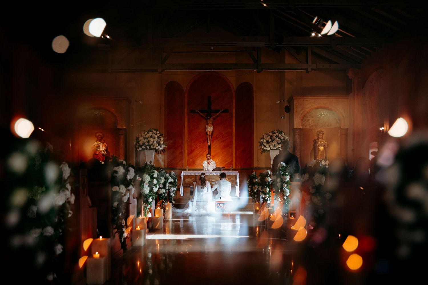 Romantic church wedding ceremony with floral arrangements, candlelight, and a crucifix altar in a warmly lit chapel.