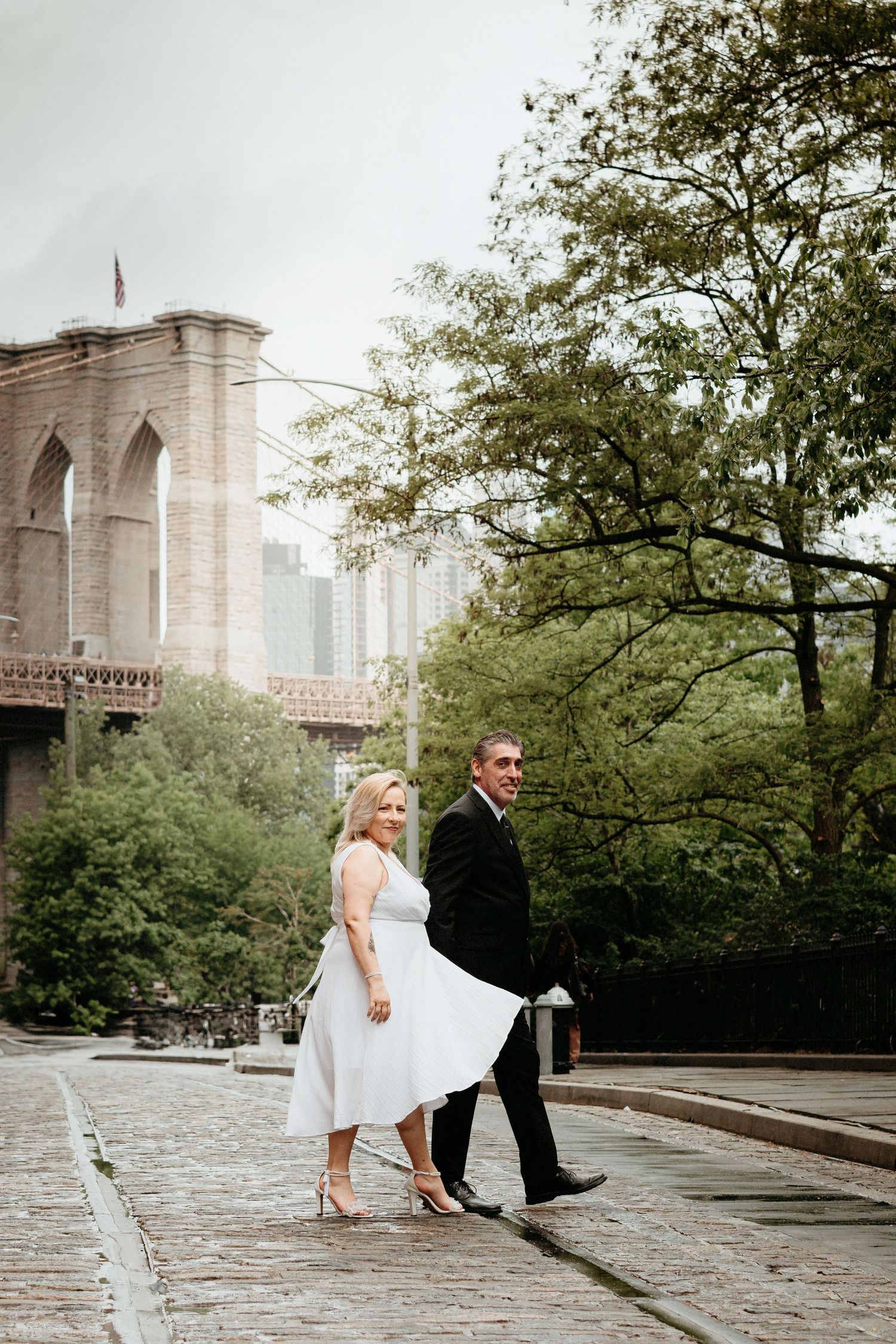 A couple walking together along the Brooklyn Bridge promenade in wedding attire.