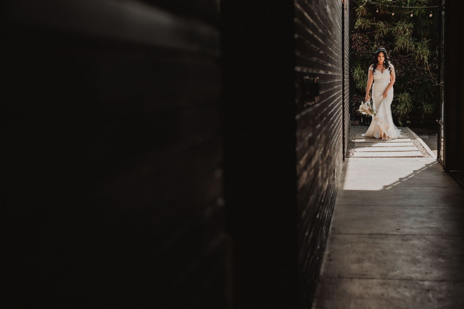 Bride in white gown stands framed by dark brick walls in dramatic light and shadow wedding portrait.