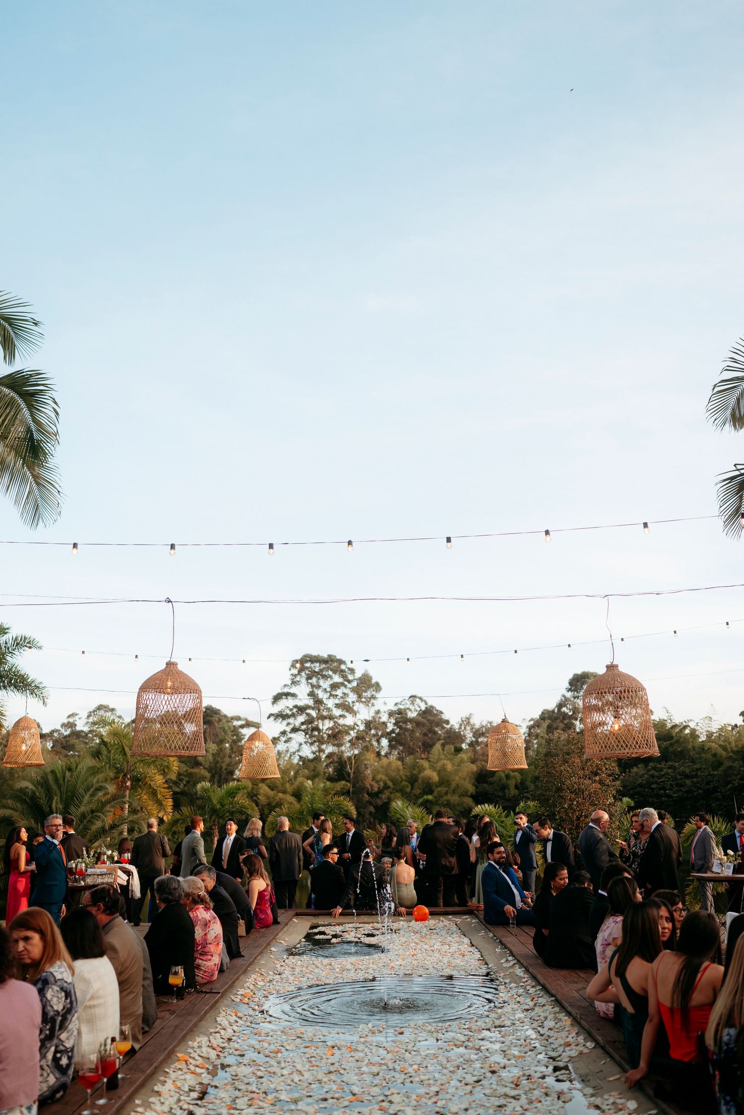 Outdoor wedding reception with string lights, wicker pendant lamps, fountain centerpiece, and guests mingling among tropical palms at dusk.