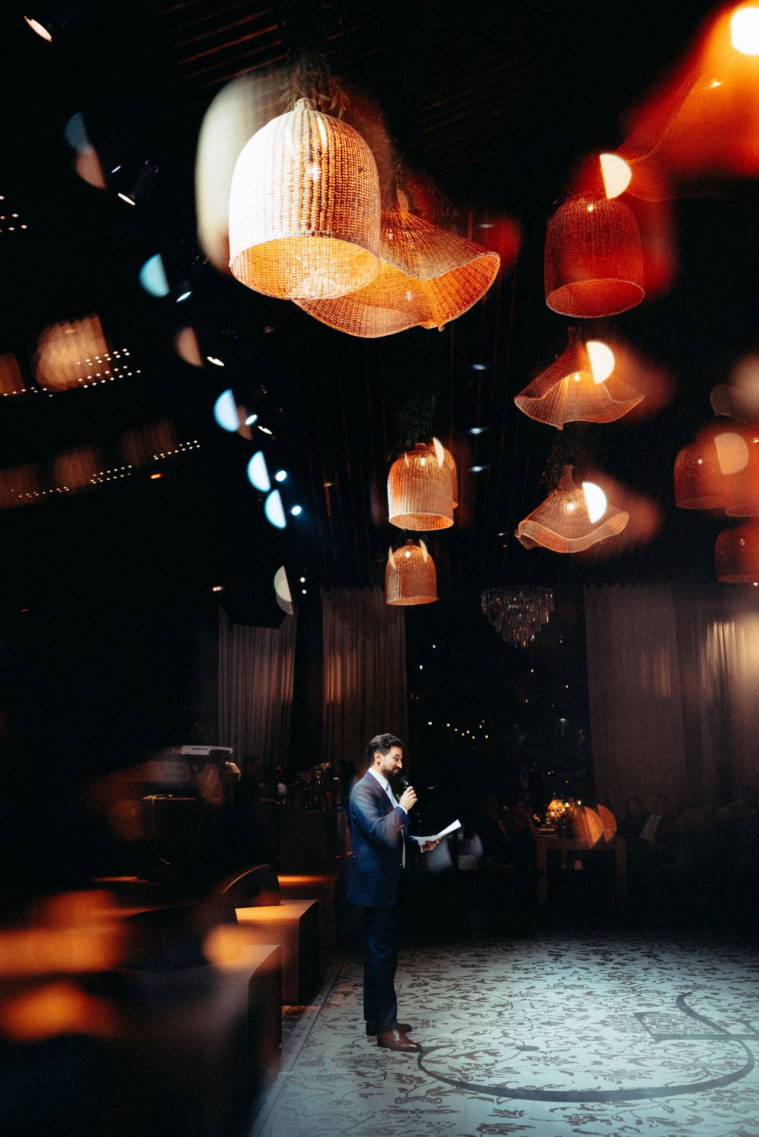 Man in suit speaking under decorative hanging pendant lights in an elegant dimly lit venue.