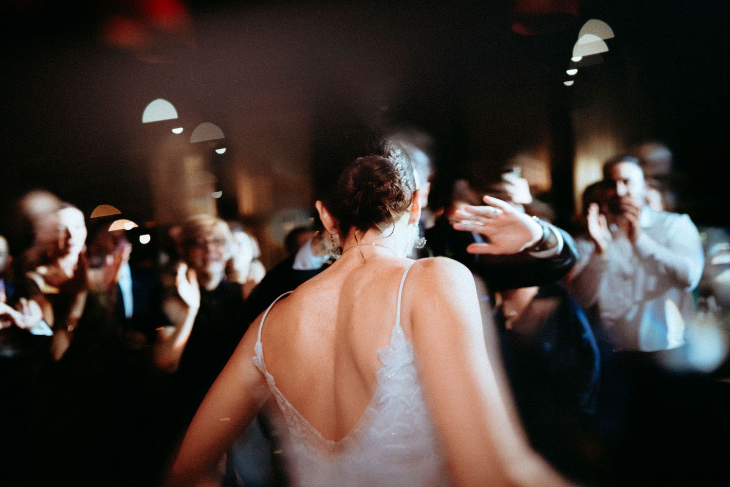 Bride in white gown dancing at wedding reception surrounded by cheering guests in dimly lit venue.
