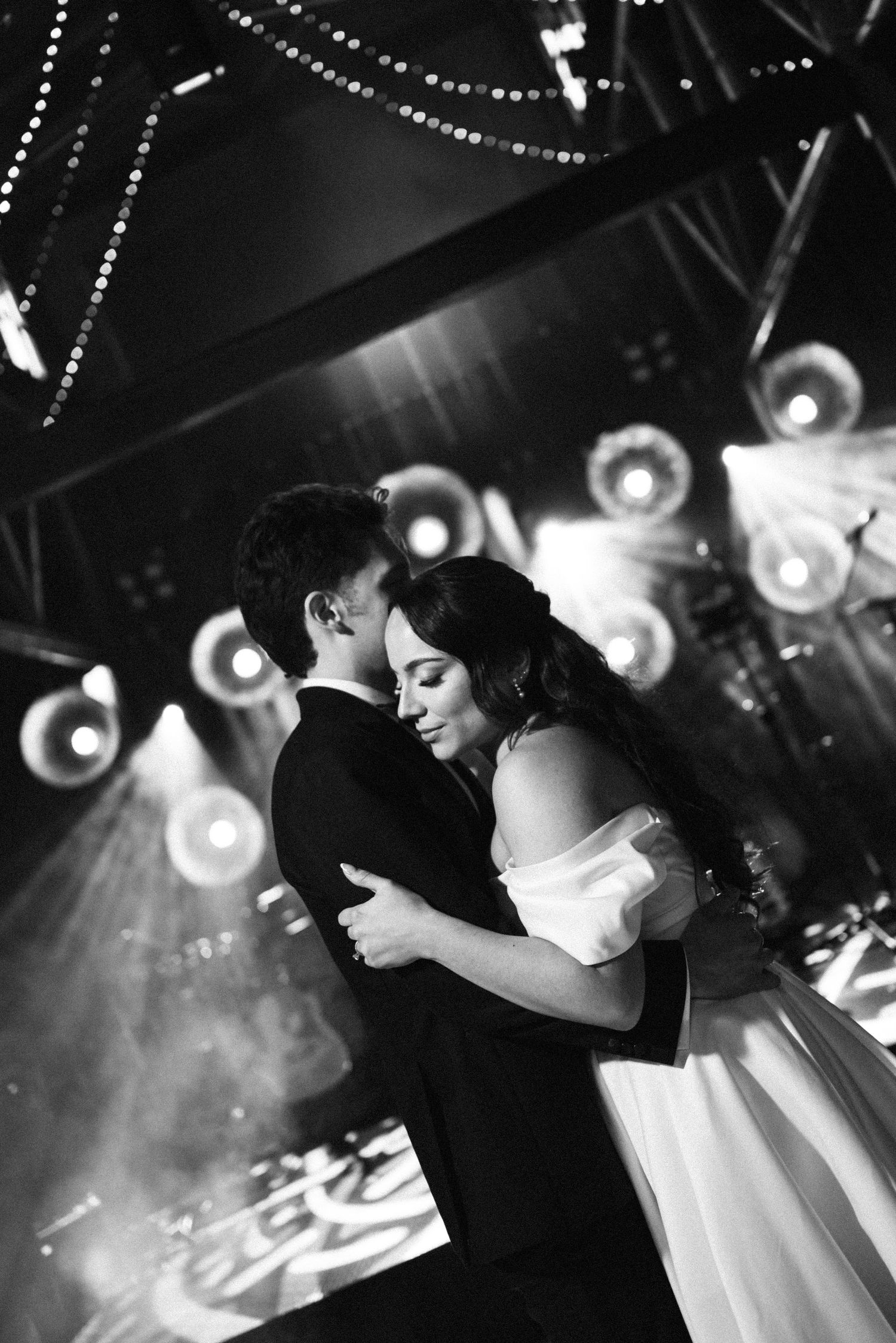 Black and white photo of newlyweds sharing first dance embrace under dramatic stage lights and fog.