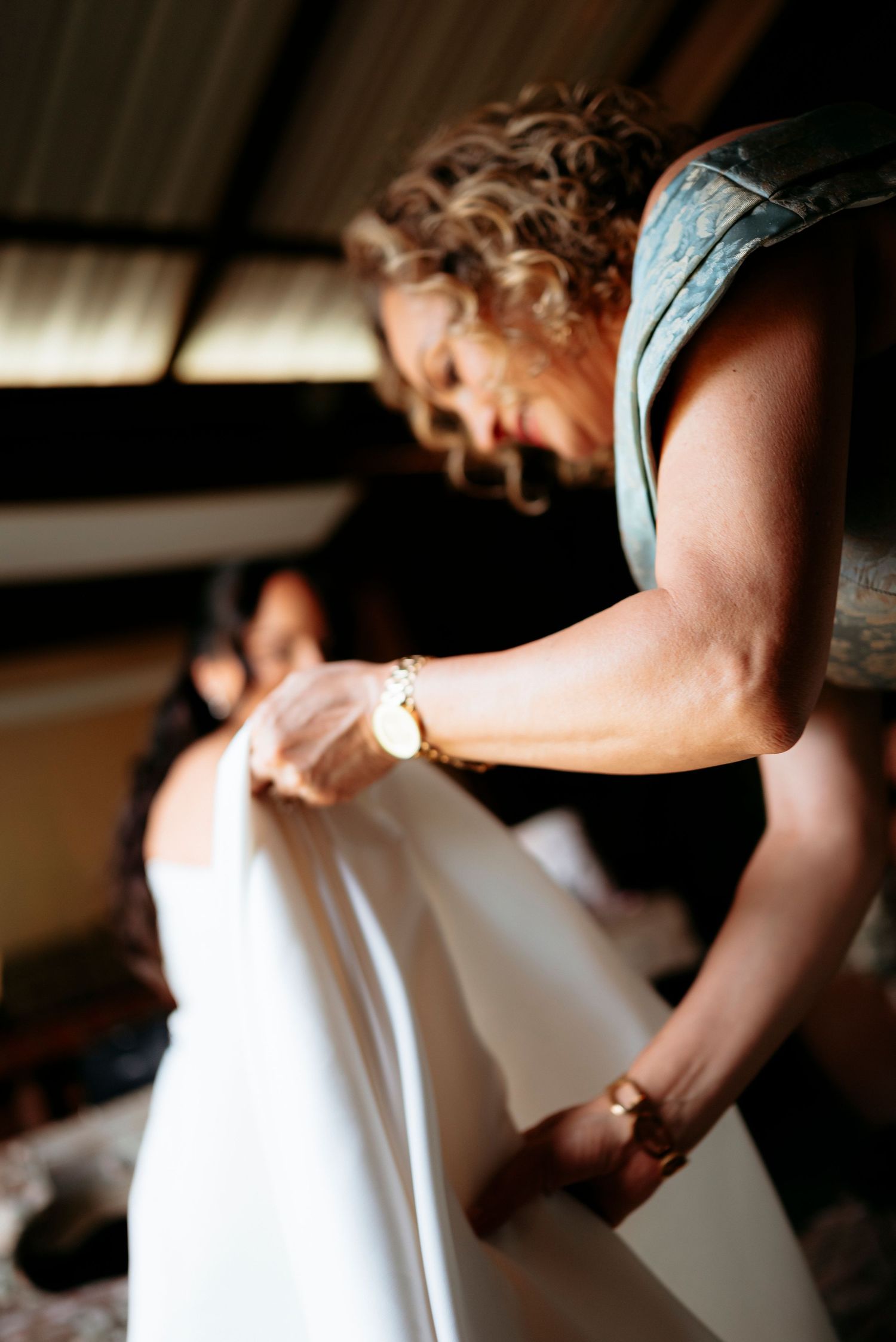 A woman in a teal dress adjusts a white wedding gown while another woman sits in the background.