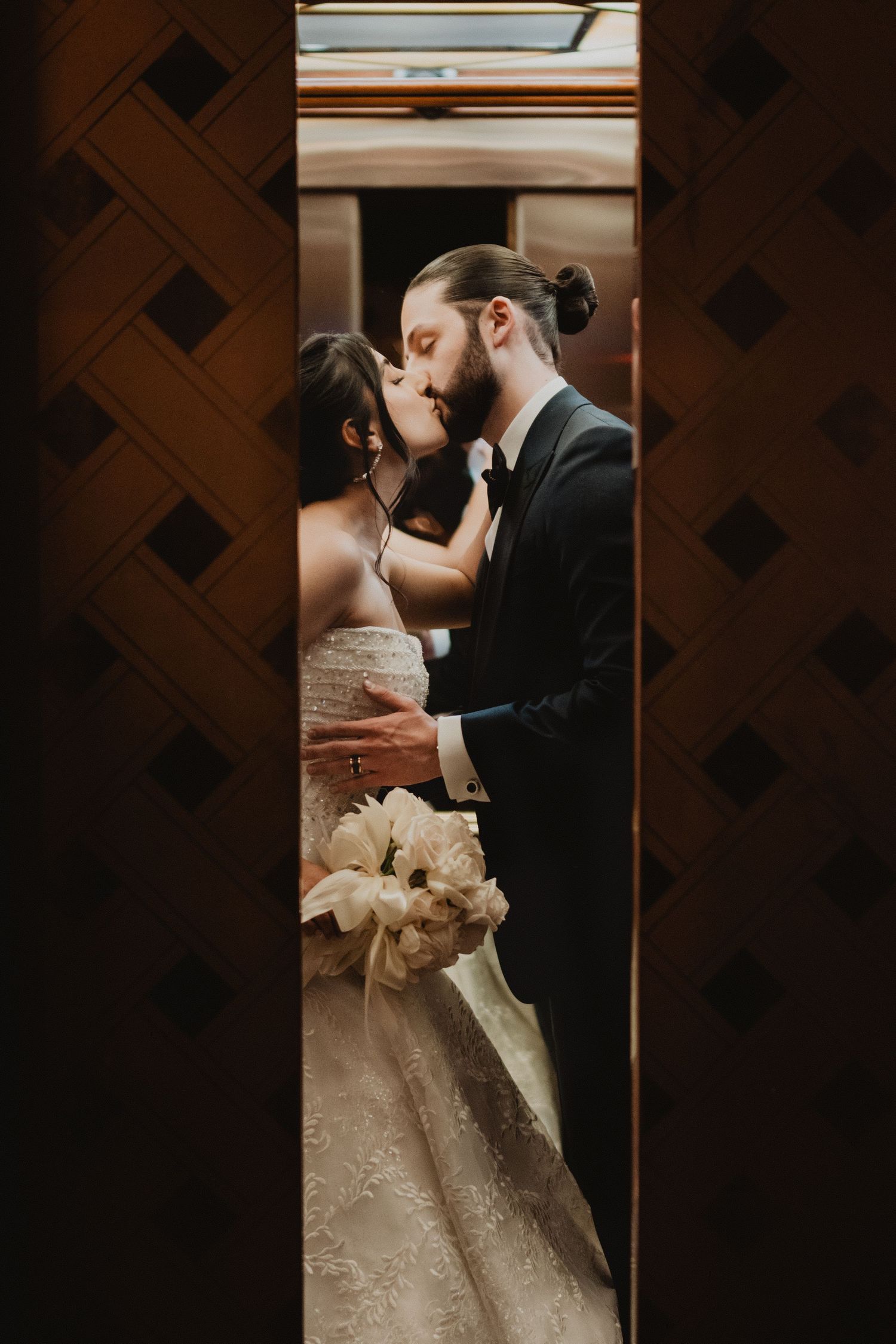 A romantic wedding moment captured through a narrow doorway showing a tender kiss and white bouquet.