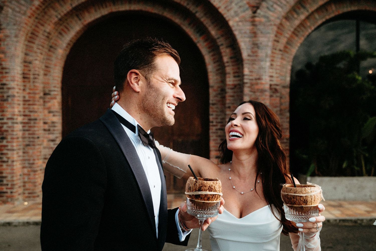 Romantic couple in formal attire shares tender moments under brick archways during wedding photoshoot.