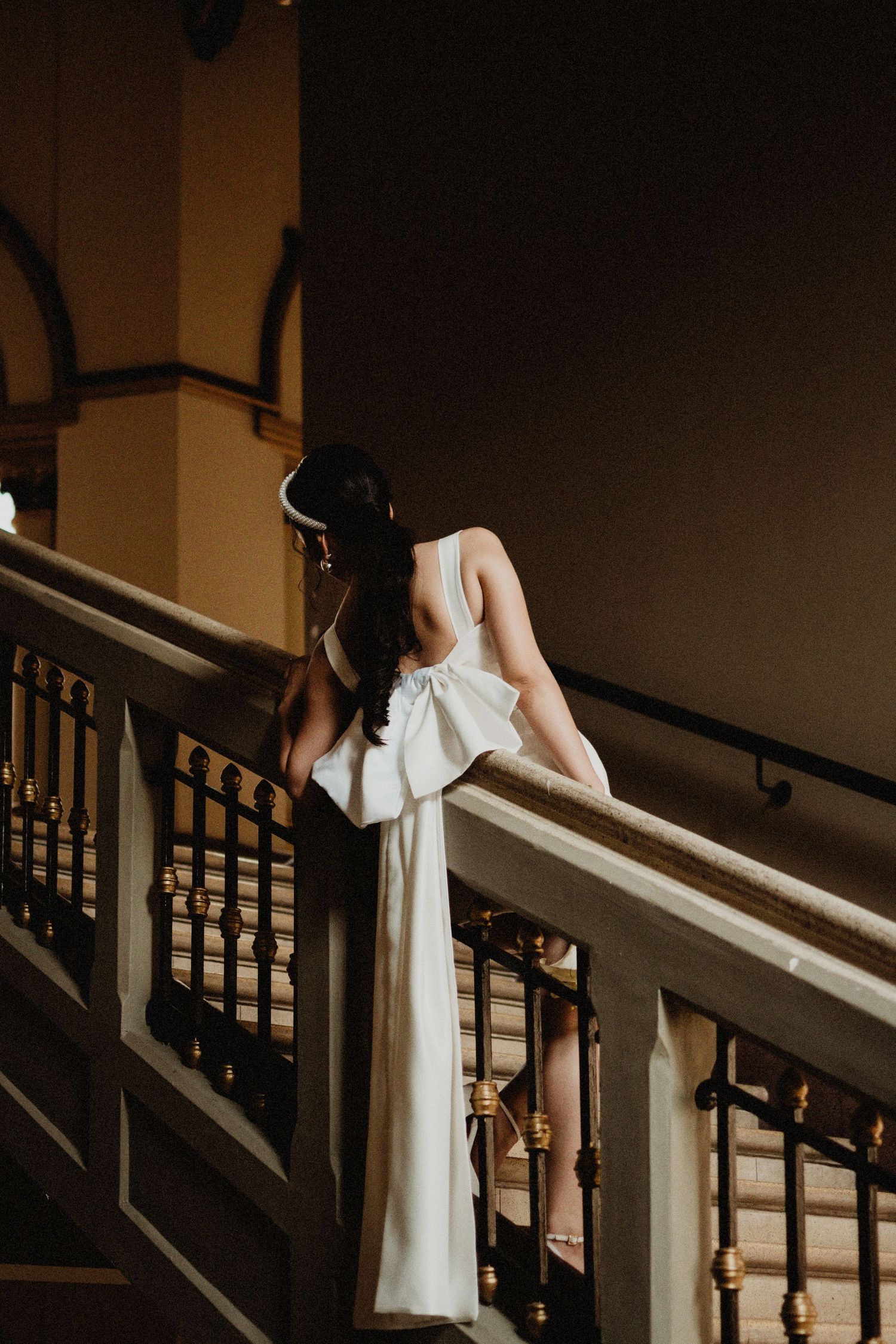 Bride in elegant white backless gown with bow detail leans on ornate staircase railing in grand venue.