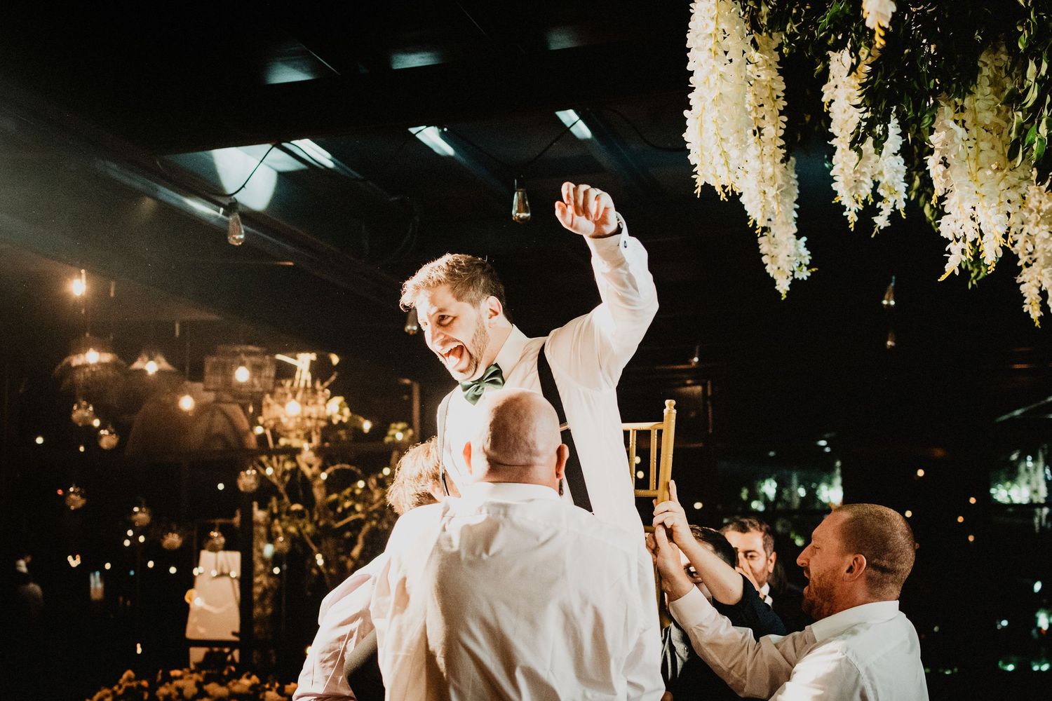 Groom lifted by groomsmen celebrating at elegant wedding reception with floral decor and warm lighting.