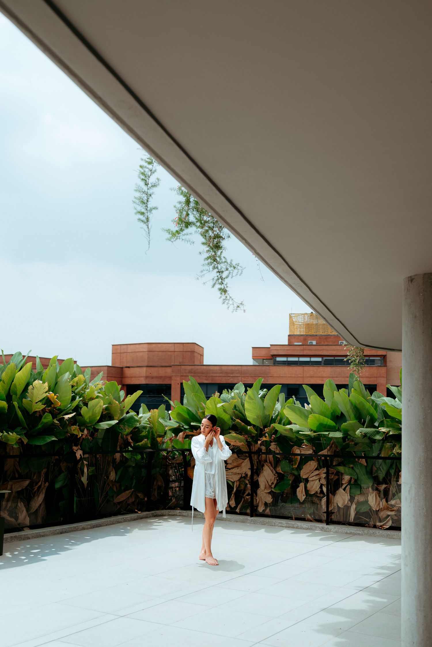 Bride in white robe standing on modern terrace with tropical plants and brick building backdrop.