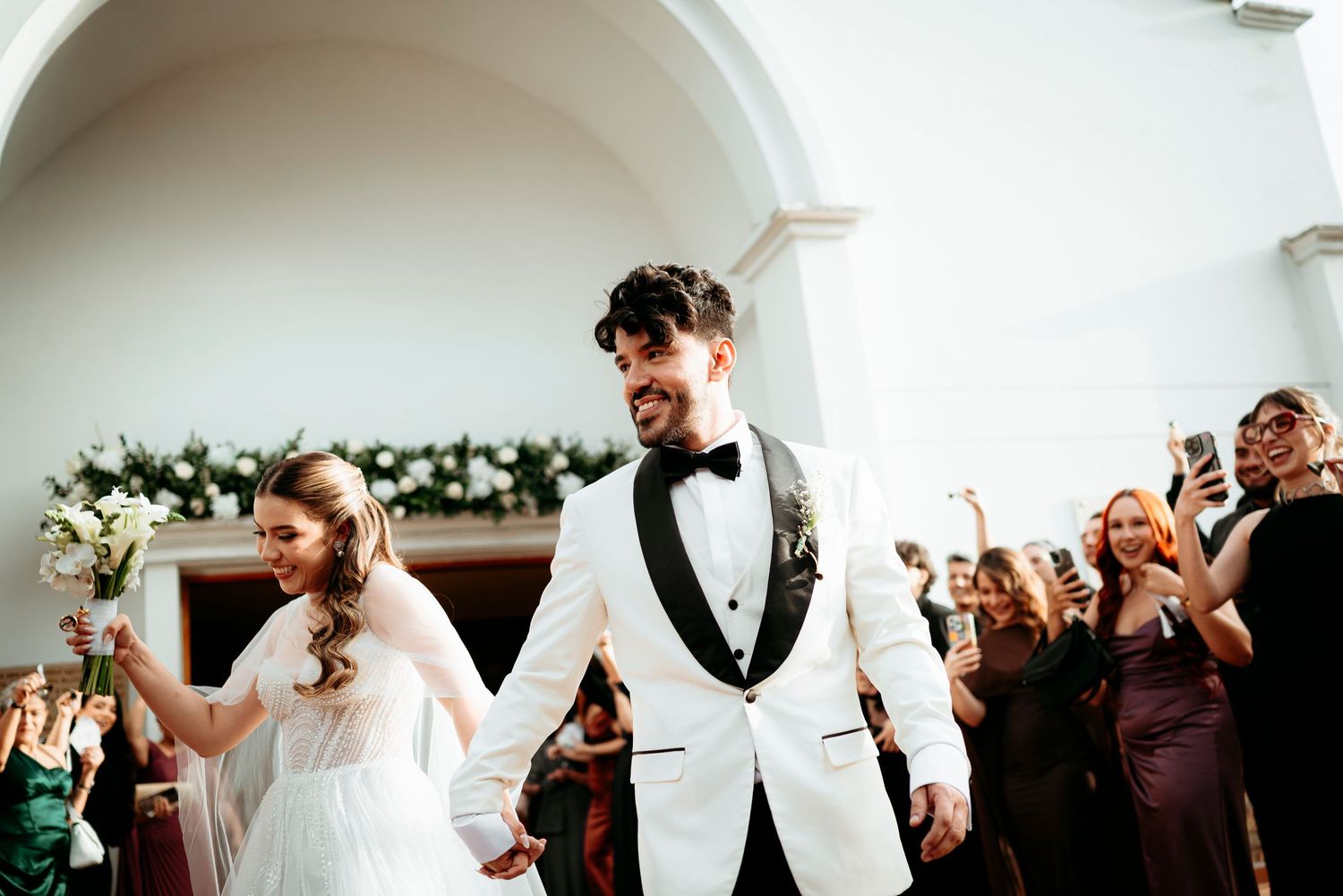 Joyful newlyweds walk down the aisle in a white church, bride holding bouquet, groom in white tuxedo.
