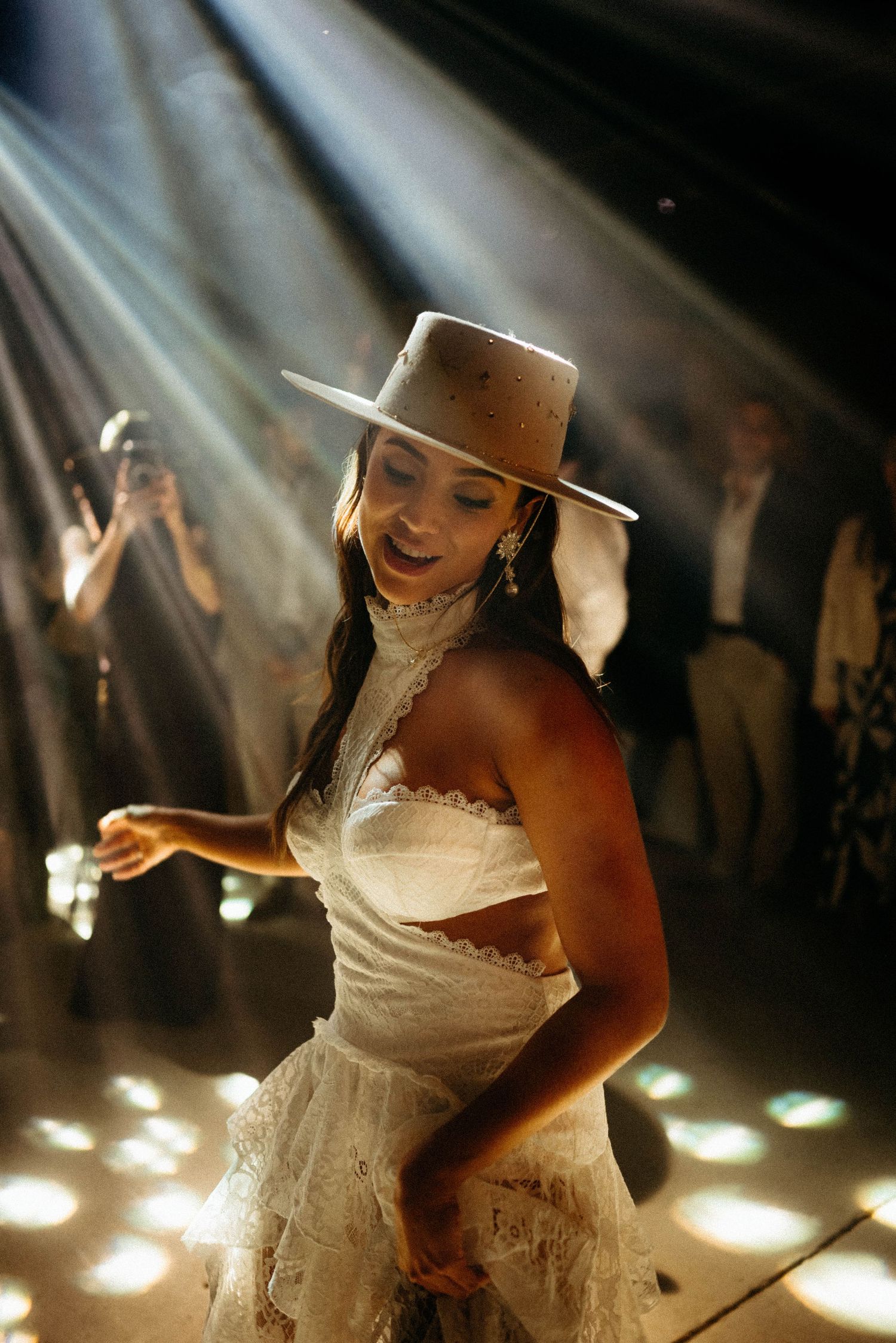 A person in a white wedding dress and brown cowboy hat dances under dramatic lighting at a reception party.