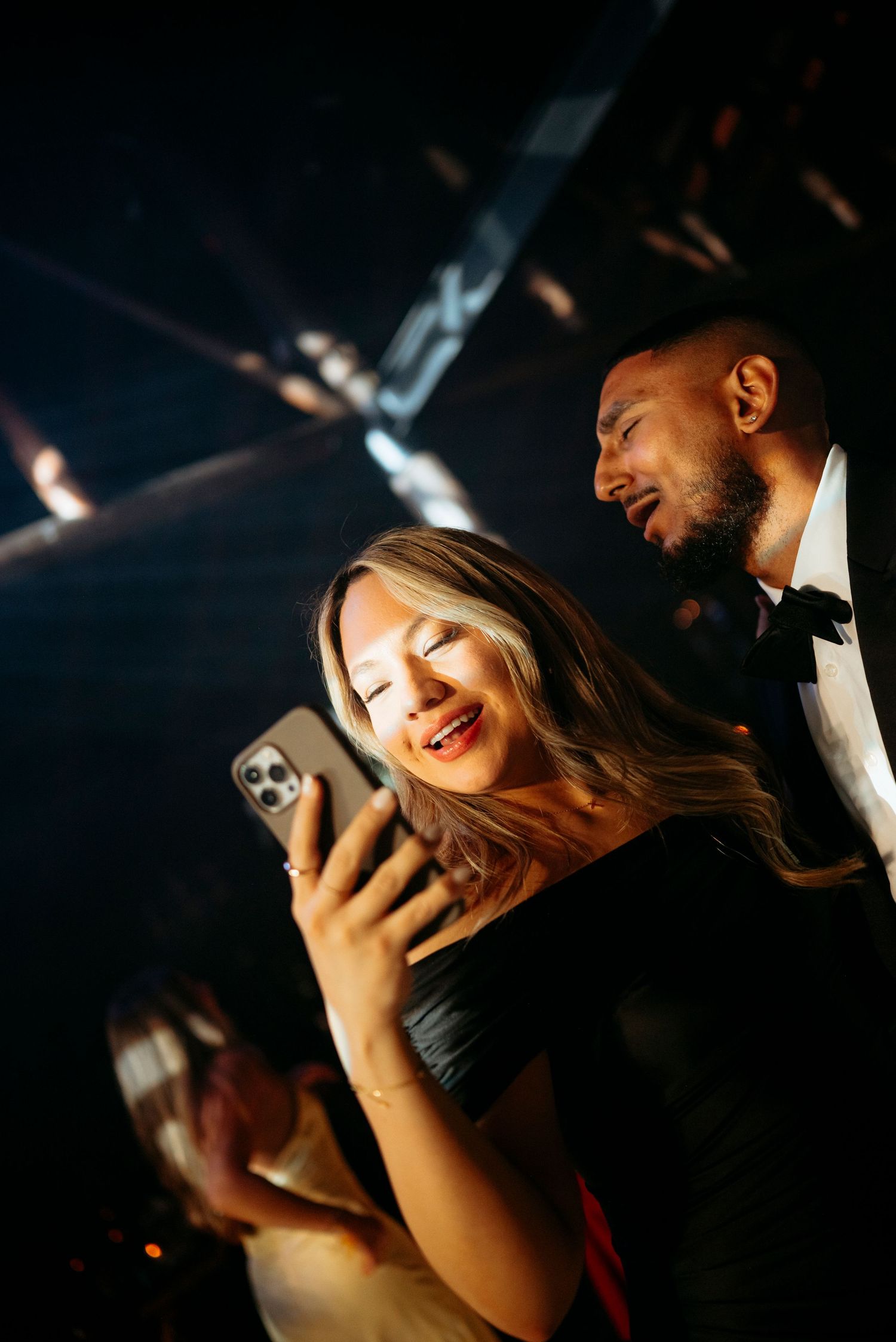 A smiling woman in a black dress views her phone while a man in a tuxedo looks on at a formal event.