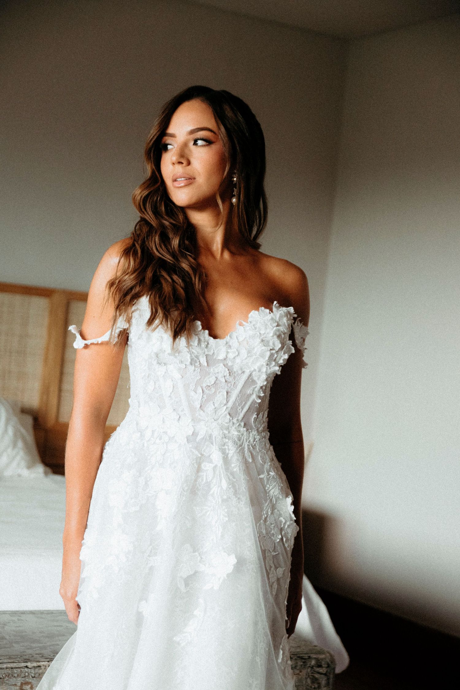 Bride in off-shoulder floral applique wedding gown with sweetheart neckline poses in softly lit bridal suite.