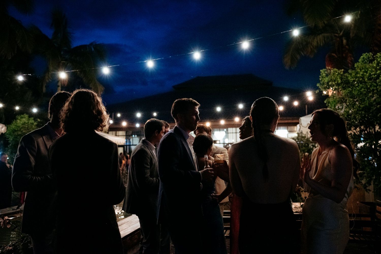 Wedding guests mingling outdoors at night under string lights surrounded by tropical palm trees.