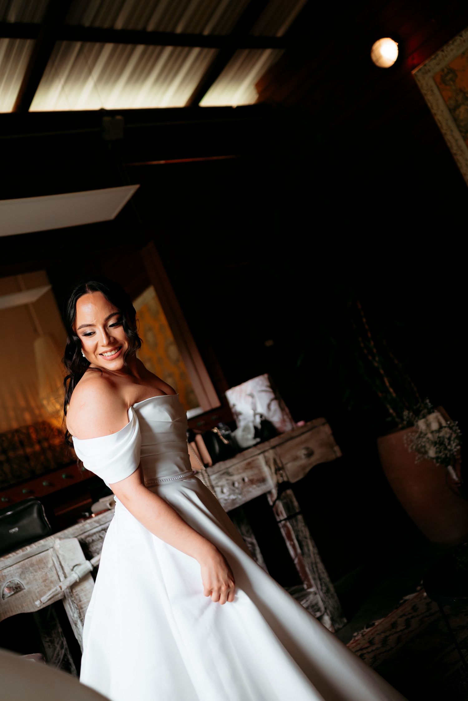 Smiling bride in an off-shoulder white wedding gown poses elegantly in a rustic indoor setting.