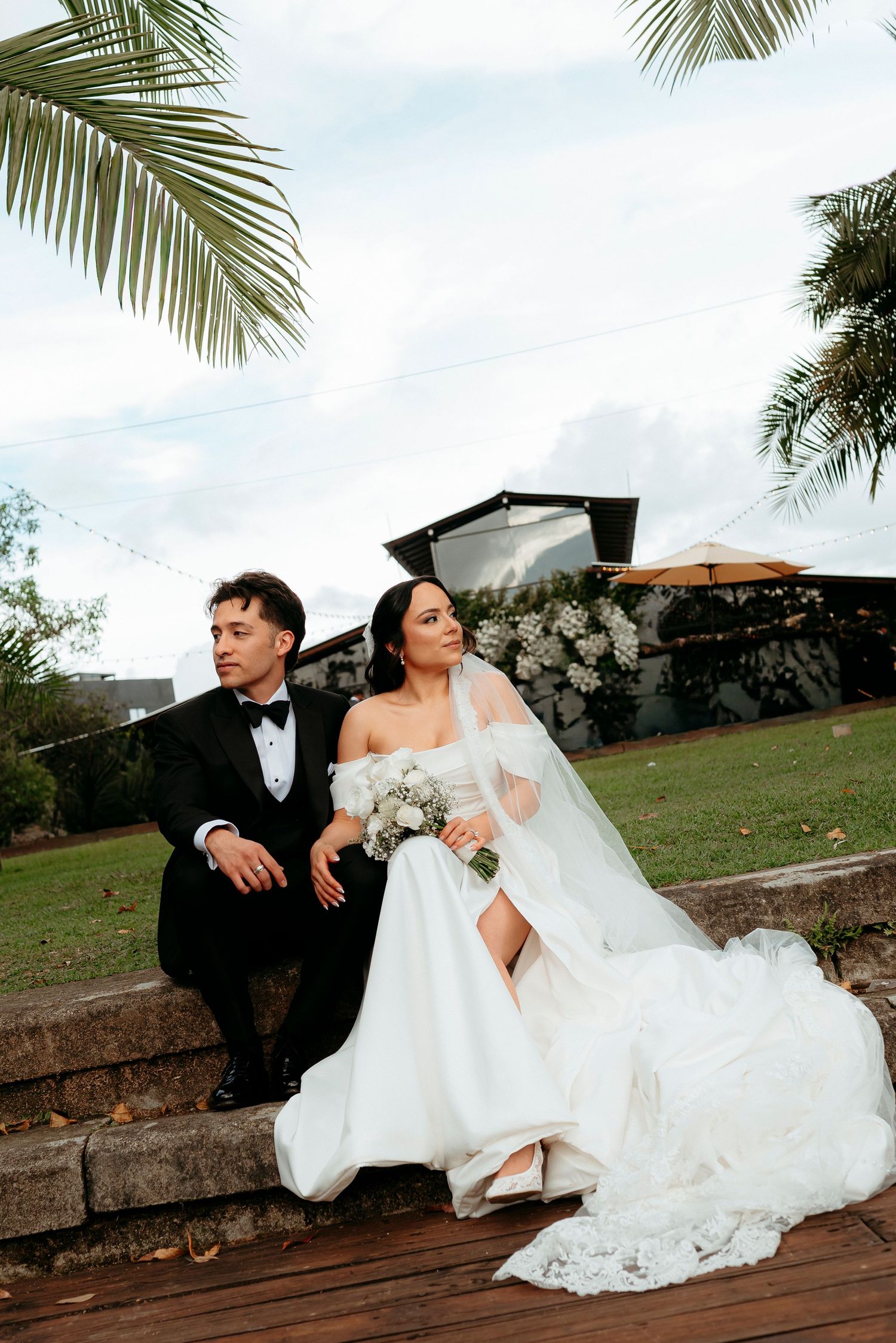 A bride and groom in elegant wedding attire sit together outdoors on stone steps, surrounded by lush greenery and white flowers.