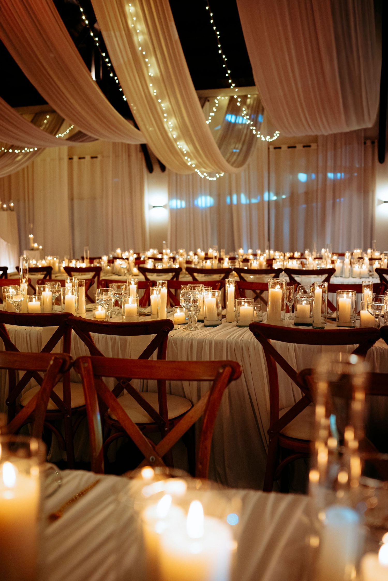 Elegant wedding reception with candlelit tables, wooden chairs, white draping, and fairy lights creating a warm romantic ambiance.