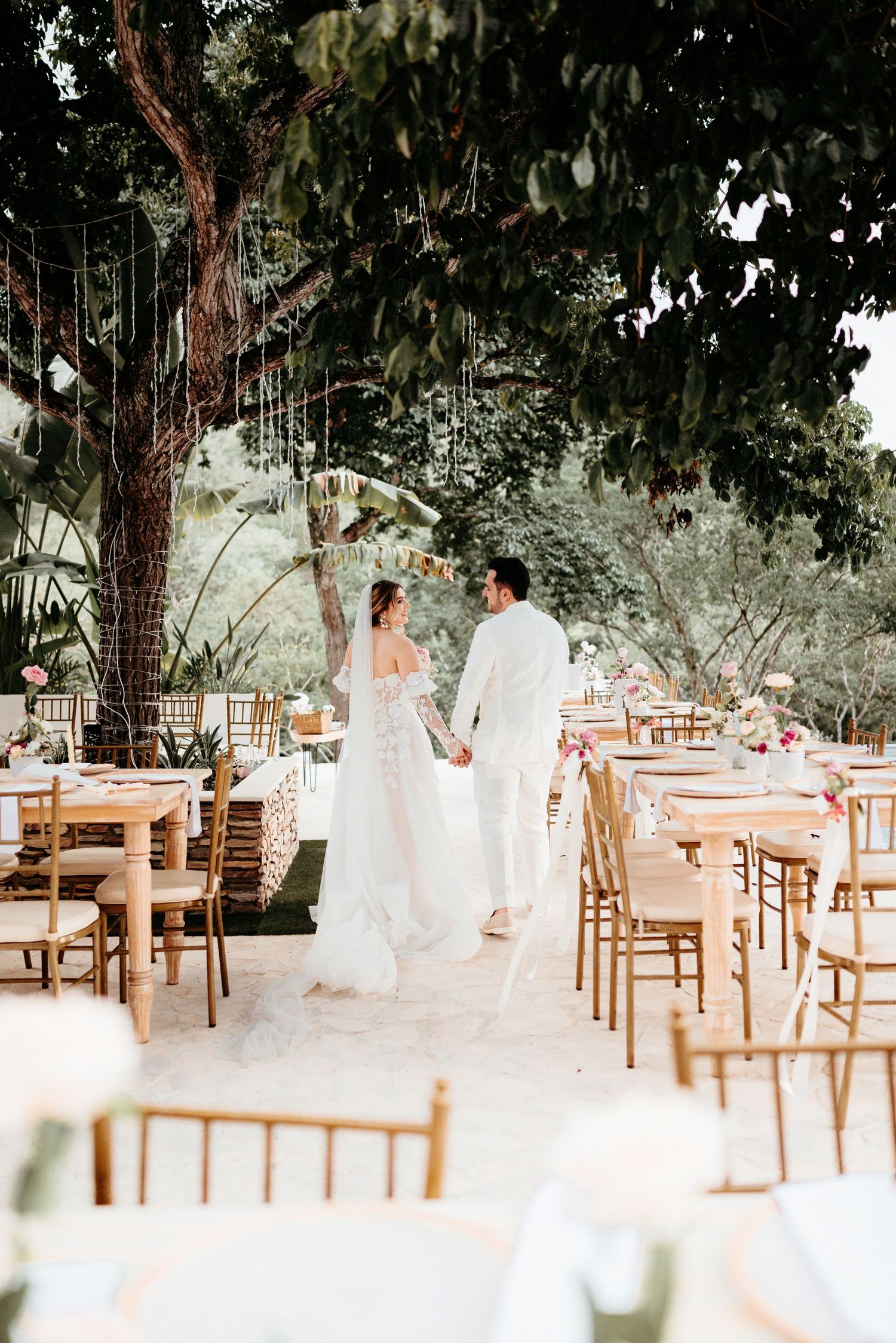An outdoor wedding reception setup with wooden chairs under a large tree.