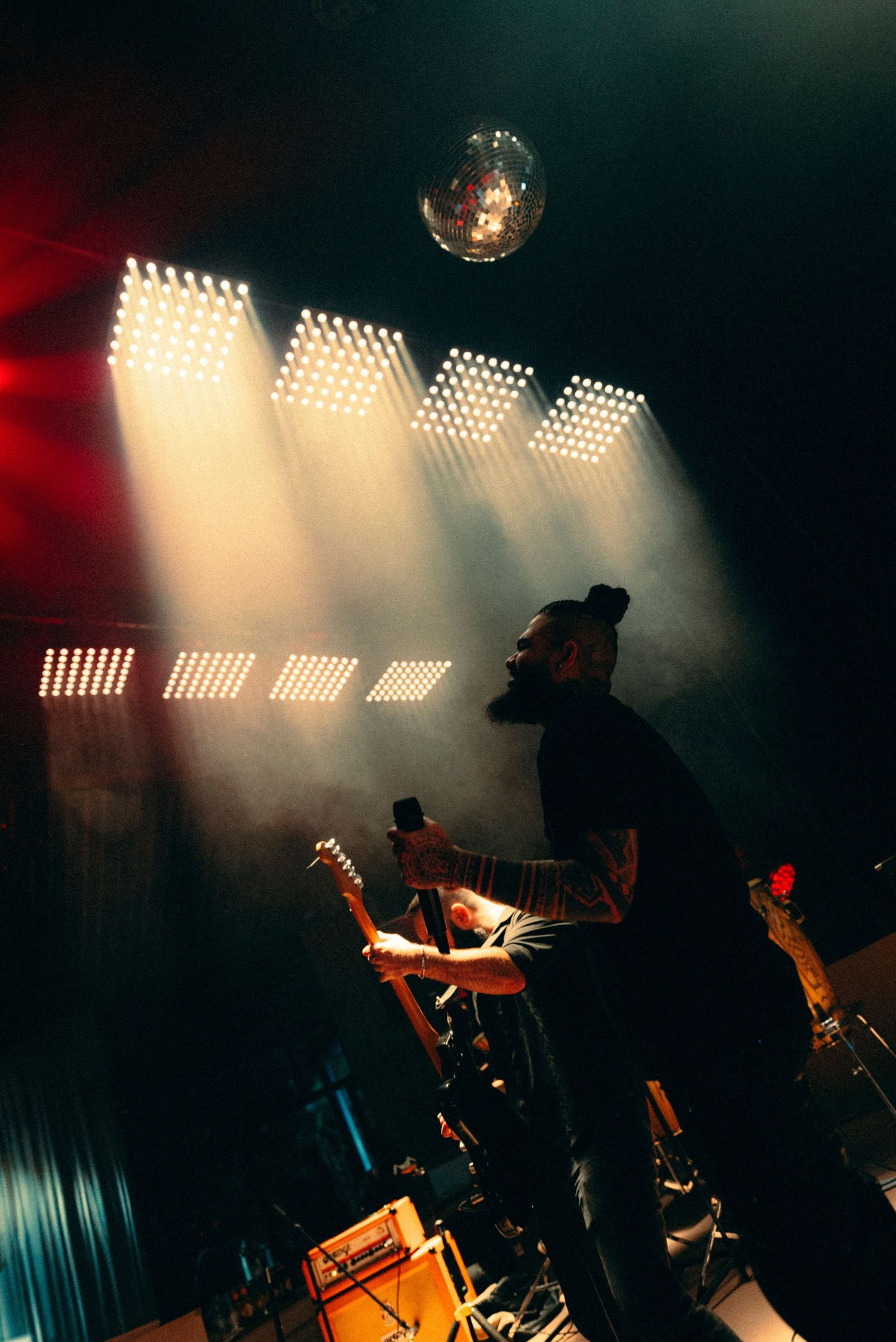 Bearded guitarist performs on a smoky concert stage under dramatic grid lights and a glowing disco ball overhead.