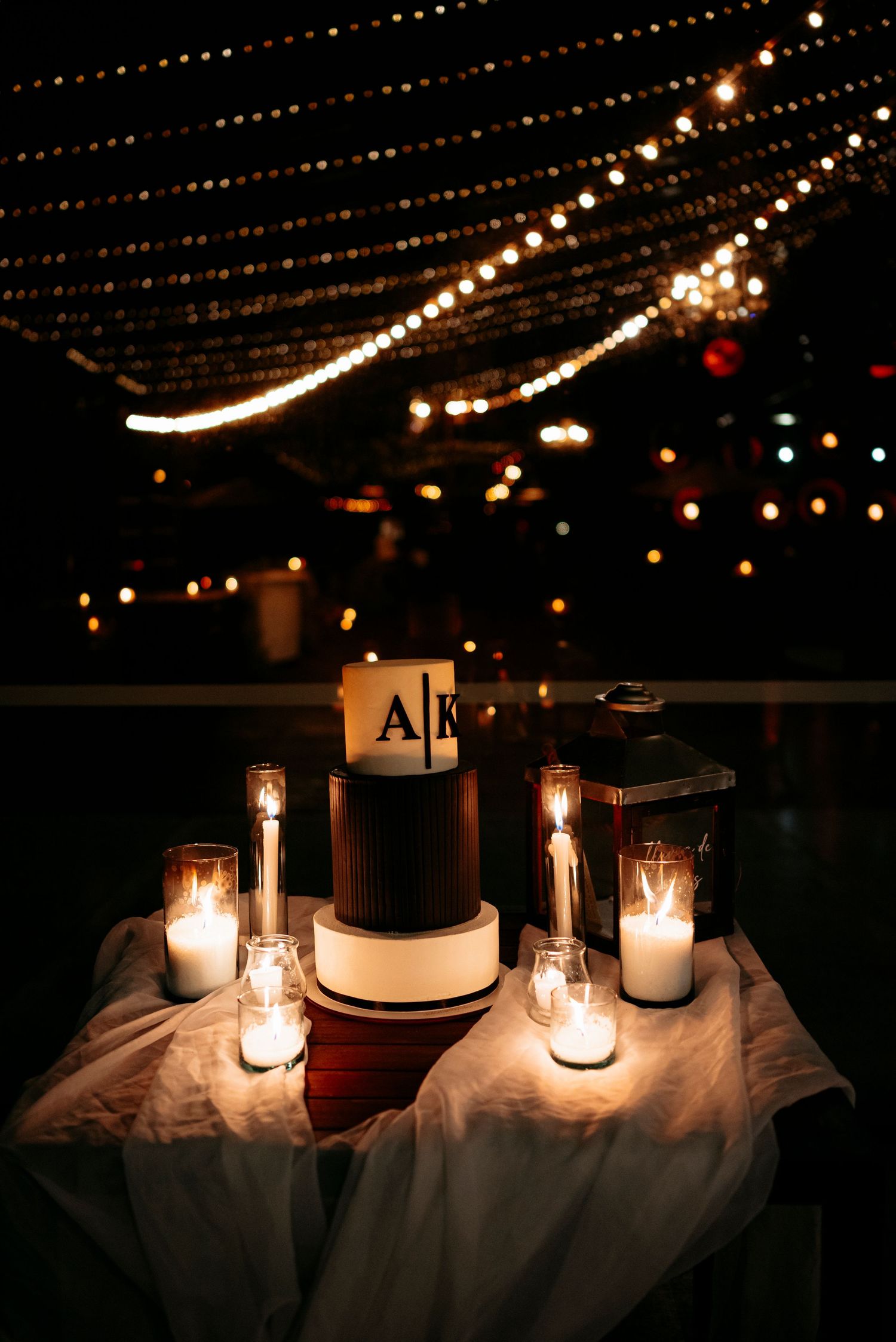 Elegant wedding cake with monogram topper surrounded by candles under string lights at night.