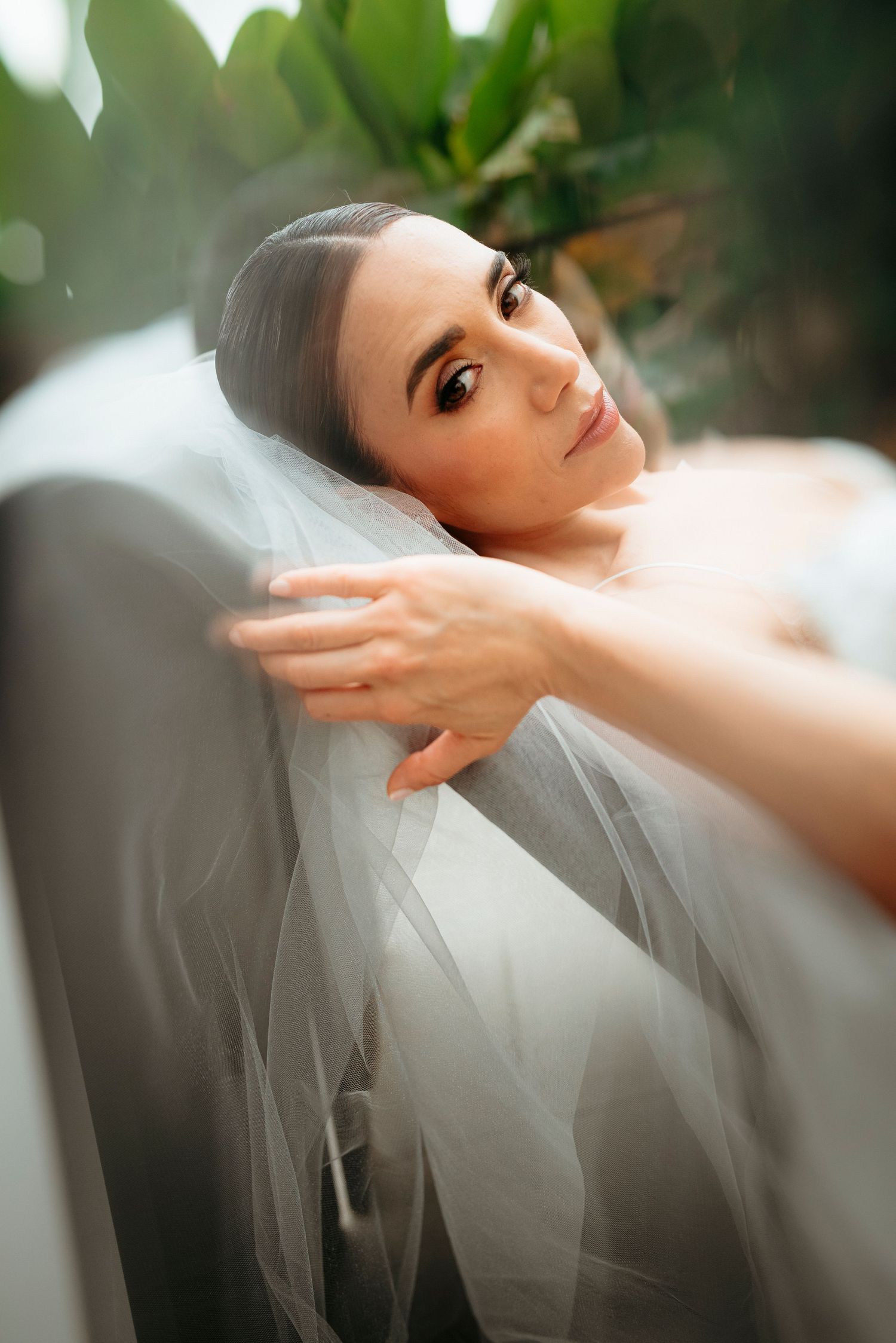 Bride draped in white veil reclines gracefully among lush green plants in soft light.