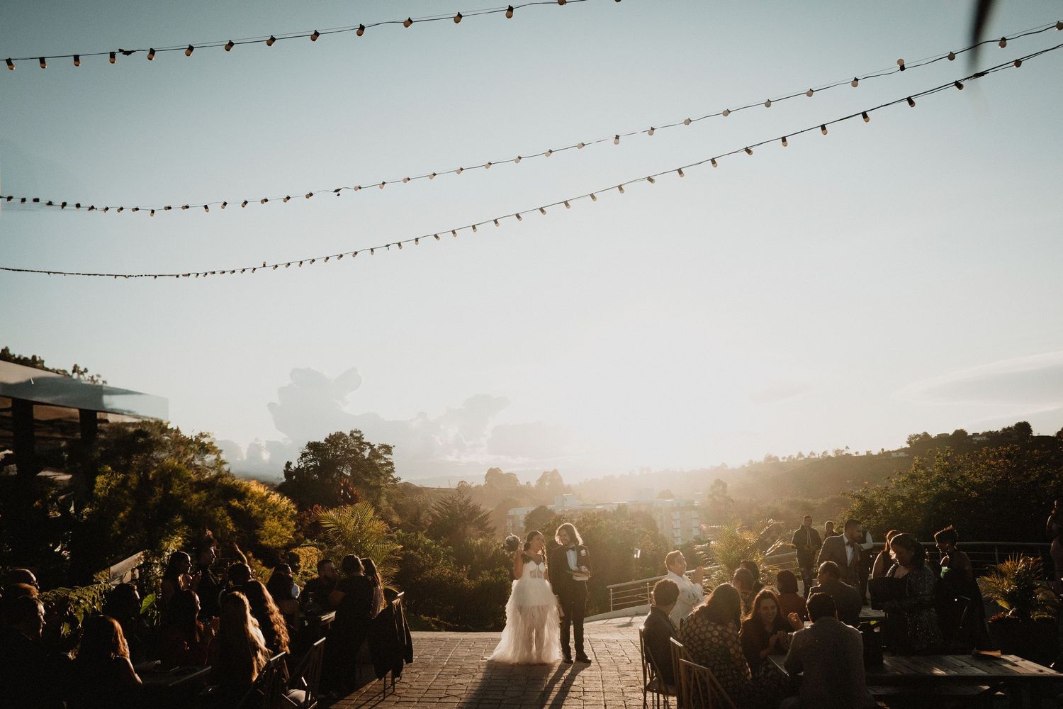 A scenic outdoor wedding ceremony under string lights at golden hour with guests watching.