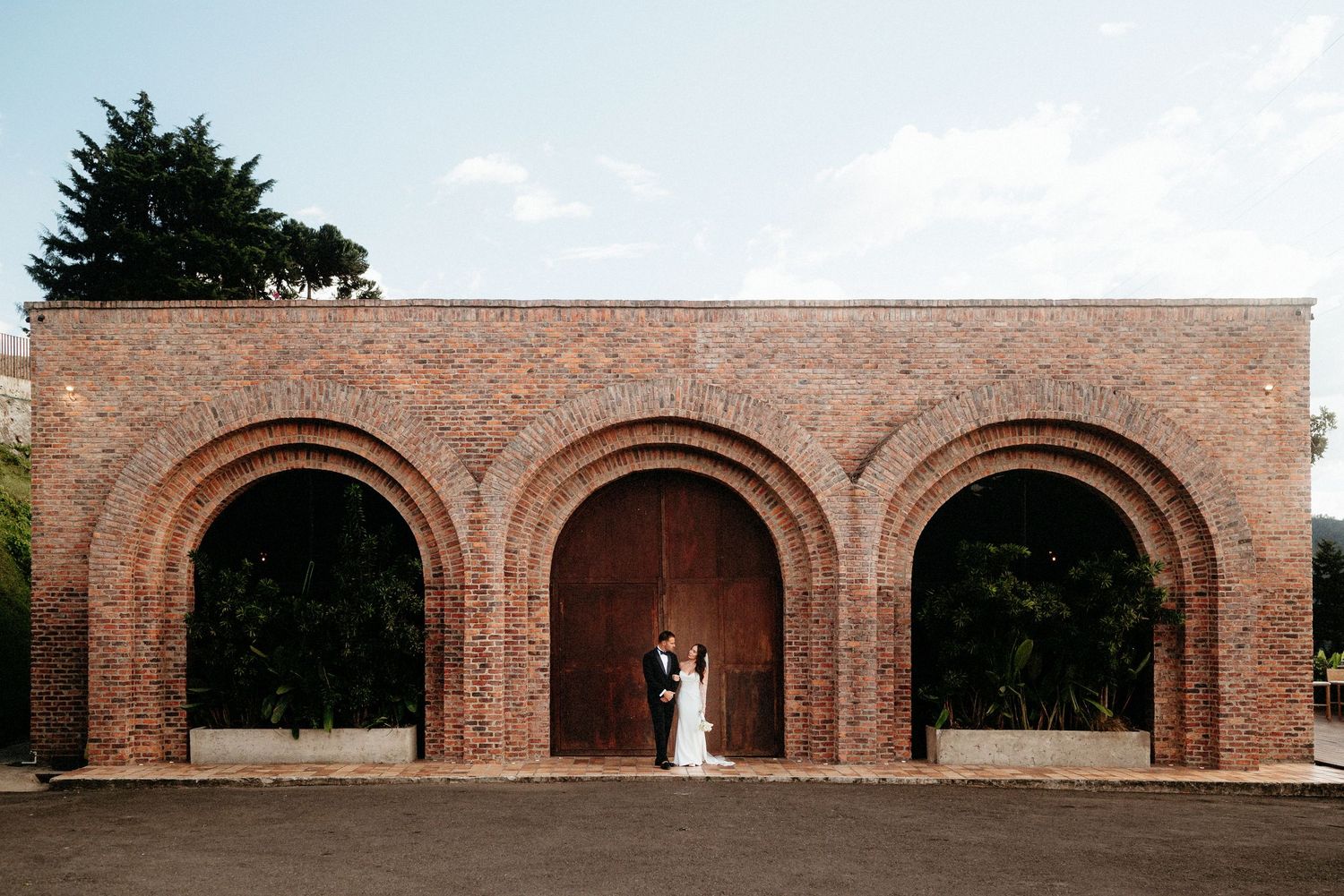 Bride and groom standing beneath brick arched wall at outdoor wedding venue on a sunny day.