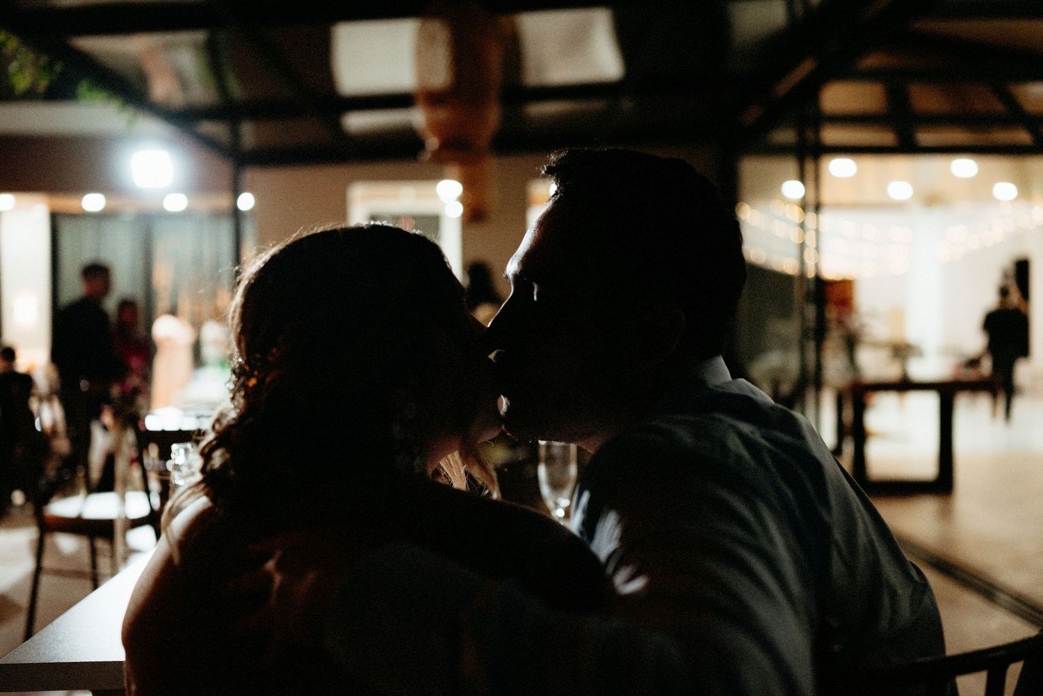 Silhouette of a couple sharing a romantic kiss at a dimly lit wedding reception venue.