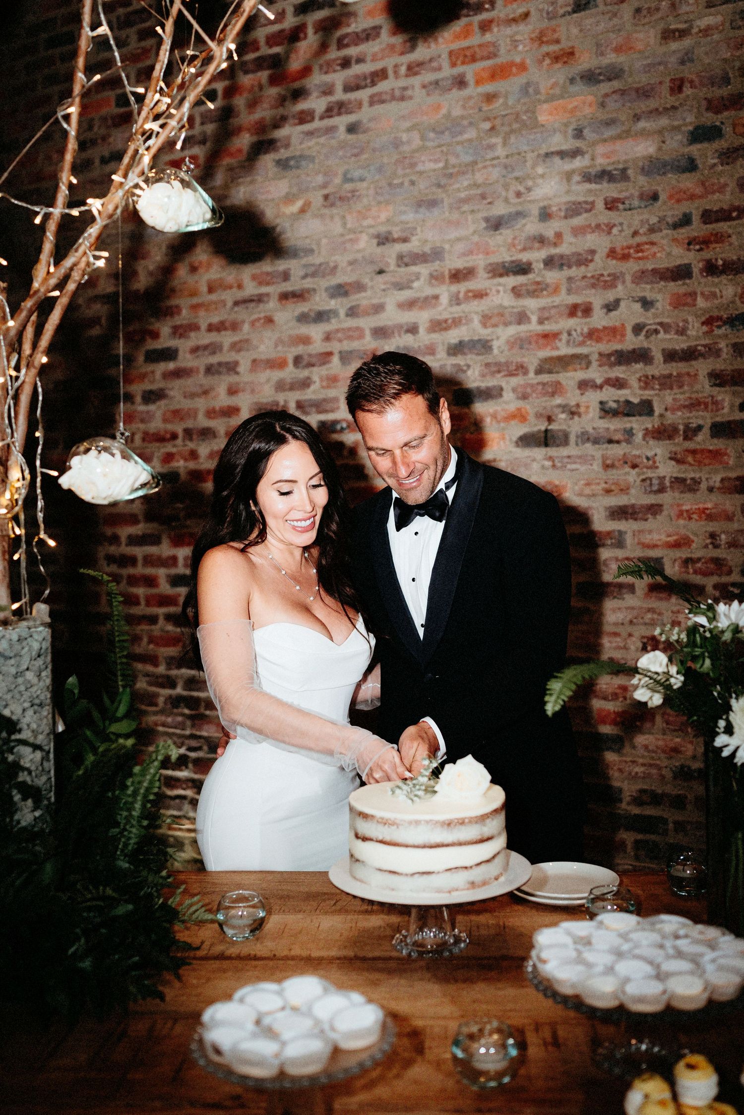 A couple in formal wedding attire cuts into a rustic naked cake at their reception with exposed brick wall backdrop.
