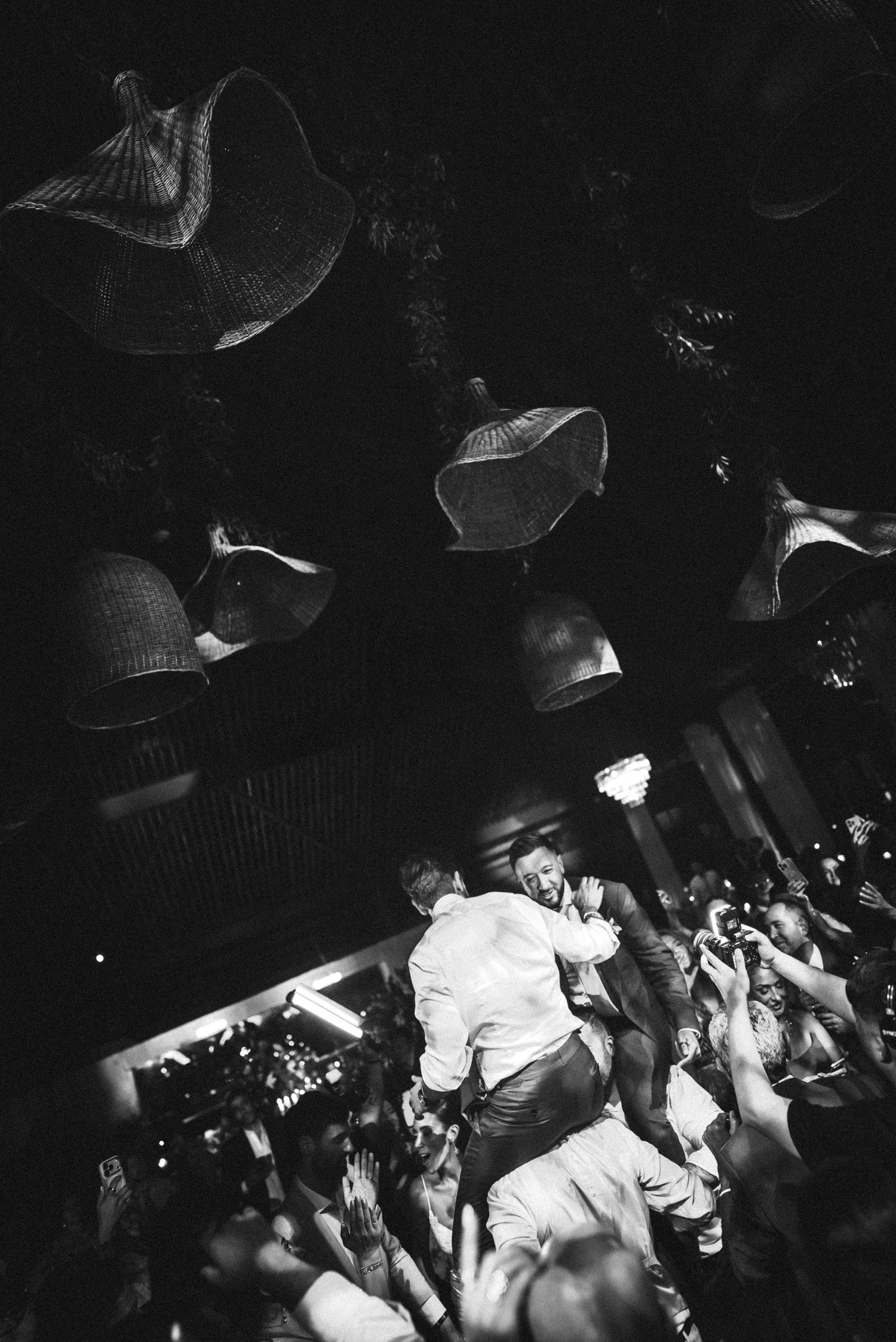 Energetic black and white crowd celebration with man lifted above guests under wicker pendant lights.