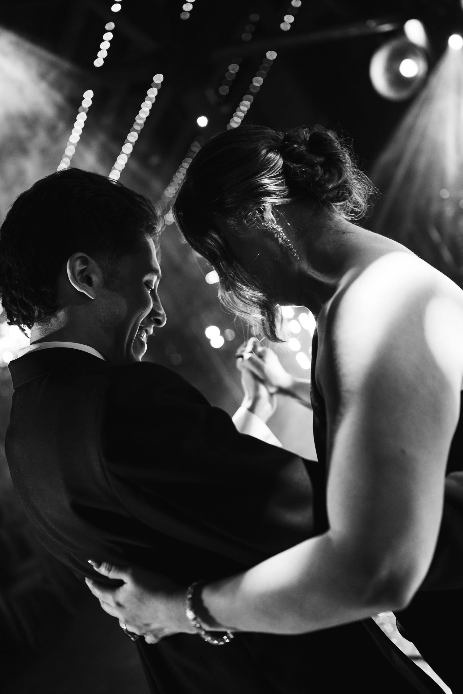 Black and white photo of a couple sharing a romantic first dance at a wedding reception under bokeh lights.
