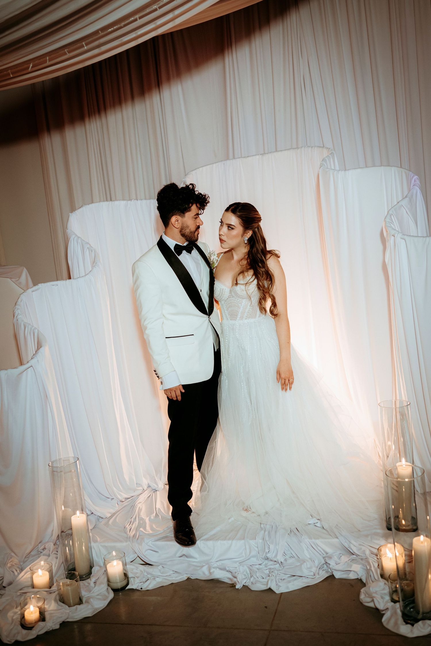 Bride in white gown and groom in white tuxedo jacket stand before draped backdrop with candles at wedding ceremony.