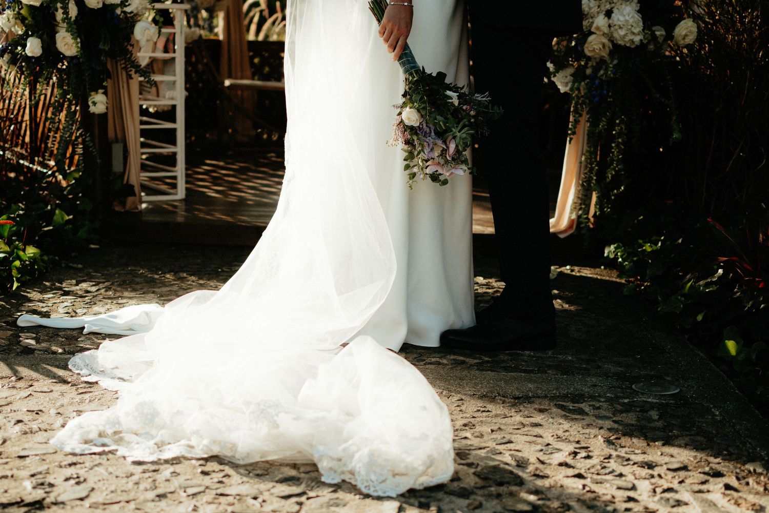 Close-up of white wedding dress train on stone path with floral and greenery details.