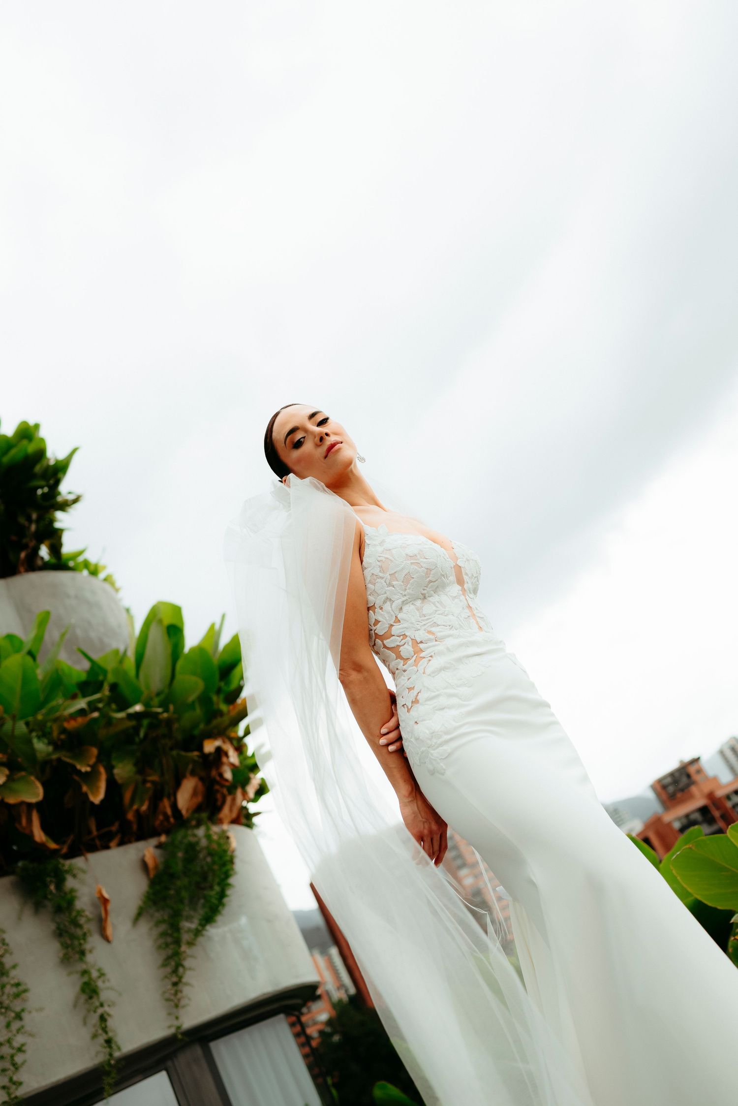 Elegant bride in lace wedding gown and veil poses on rooftop terrace surrounded by tropical greenery.
