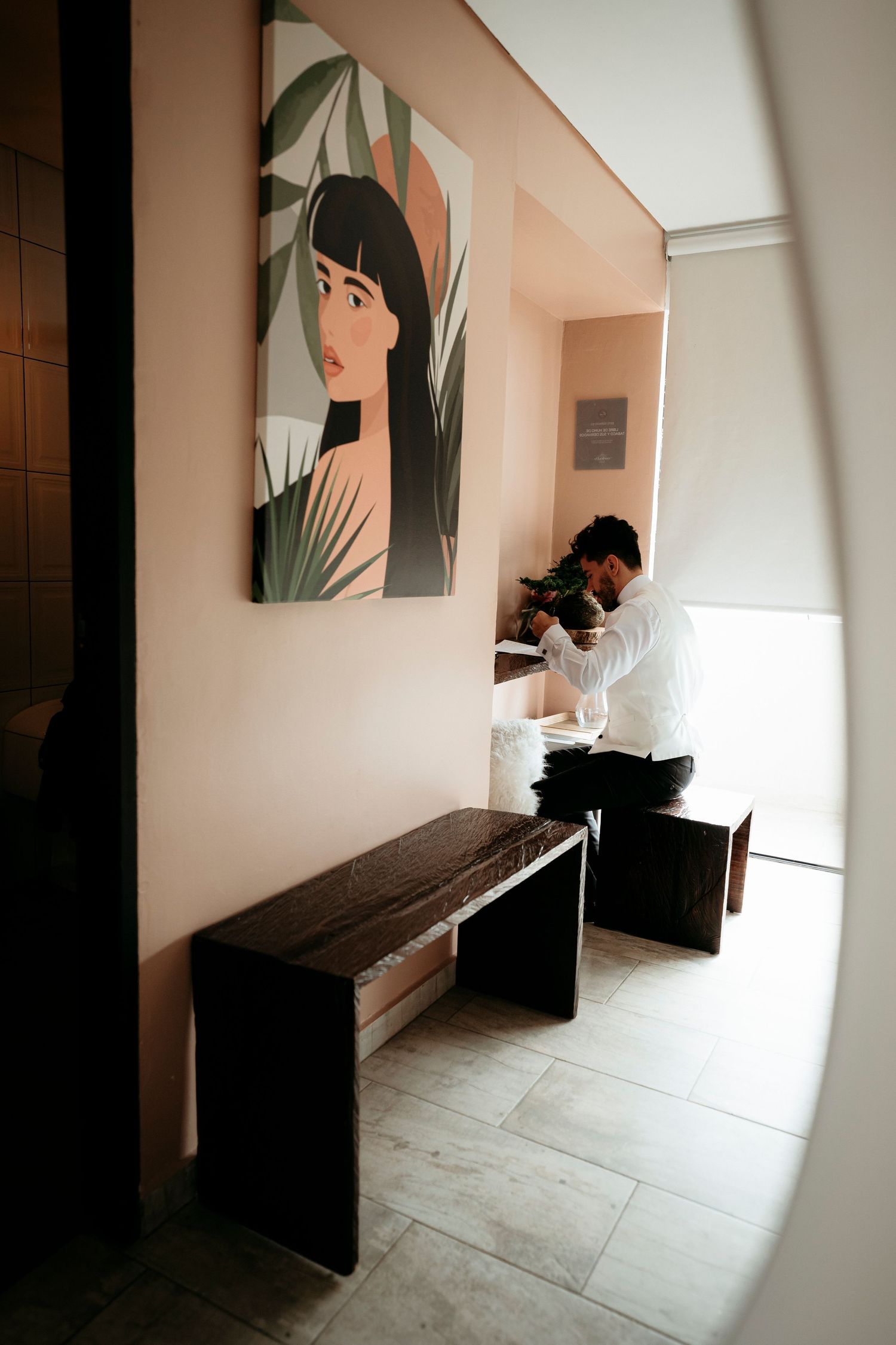 Man in white shirt arranges flowers in a modern hallway with dark wood bench and decorative portrait painting on pink wall.