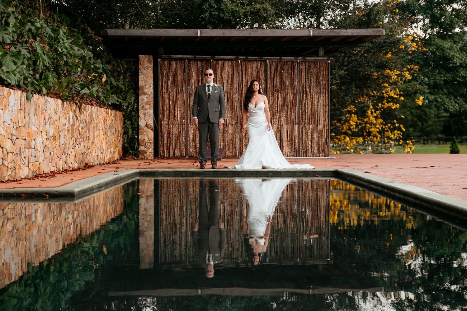 A couple in wedding attire stands by a still pool creating a perfect reflection against rustic wooden doors.