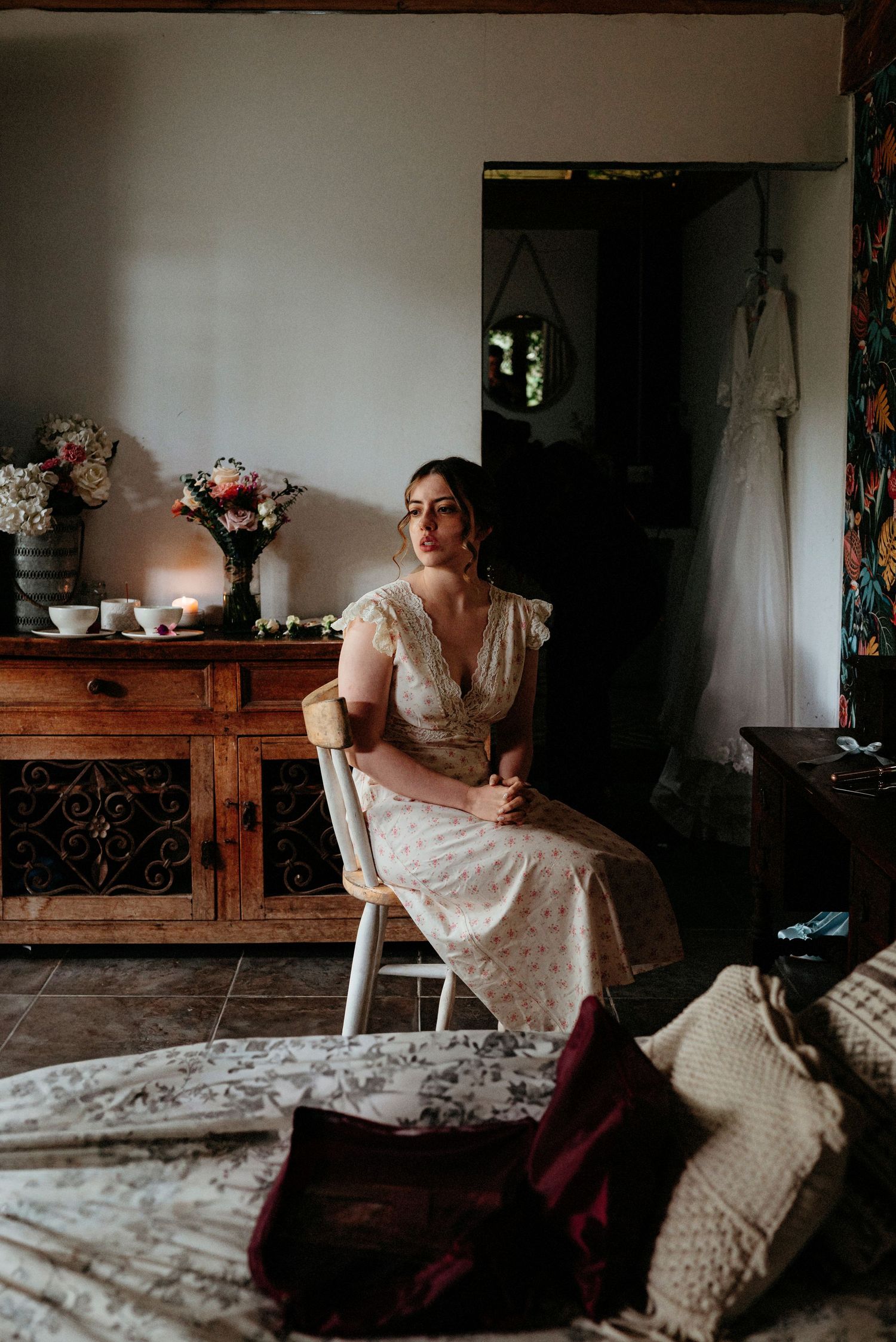 A person in a vintage-style polka dot dress sits near a wooden table with flower arrangements and a hanging dress in background.
