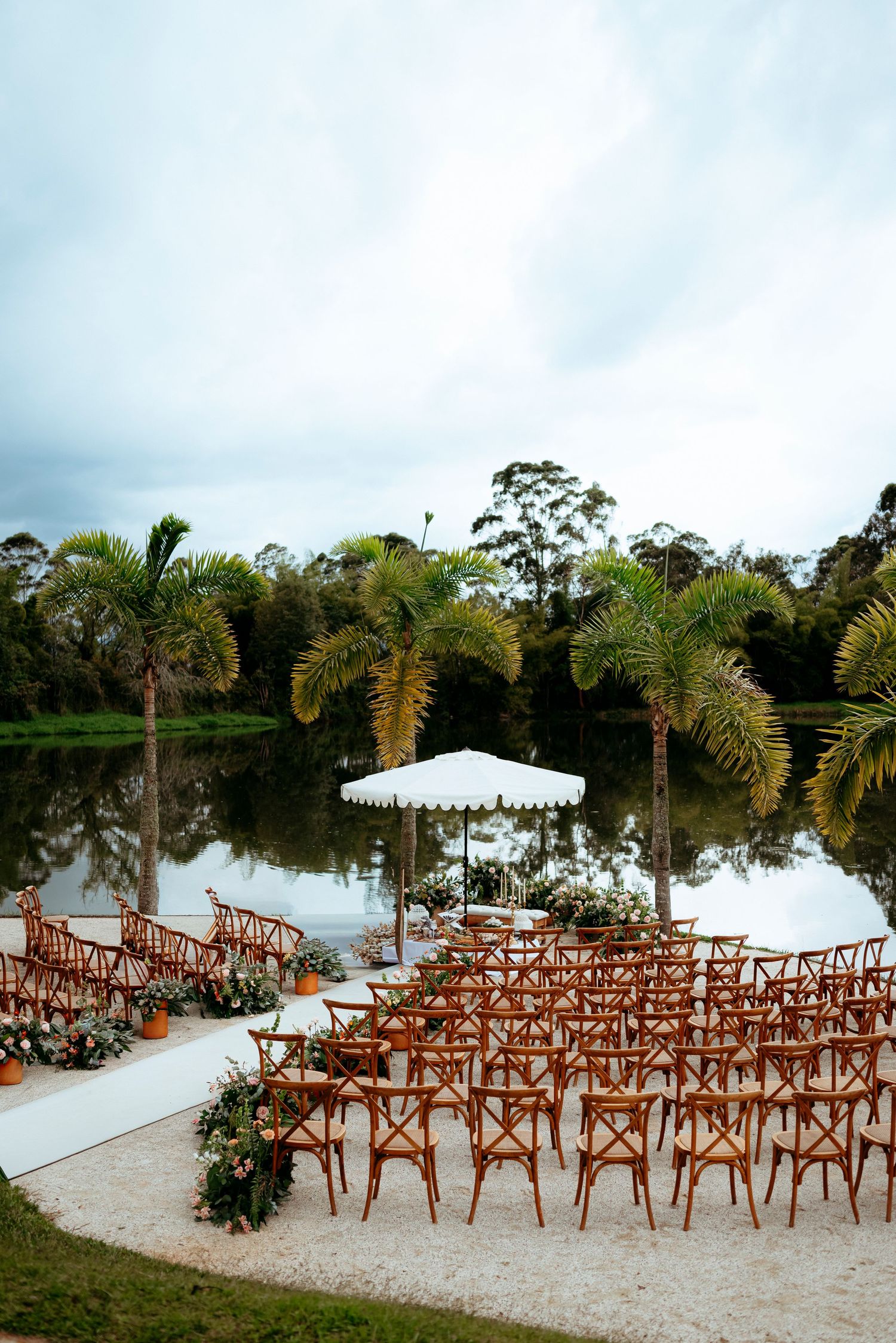 Elegant outdoor wedding ceremony setup with wooden cross-back chairs facing a white canopy altar by a serene lake surrounded by palm trees.