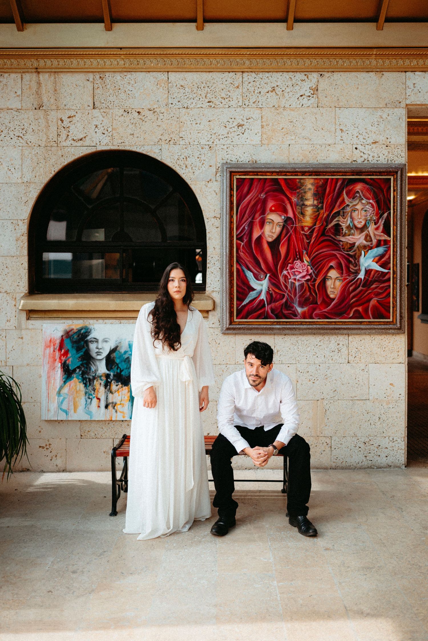 Couple posing in elegant venue with colorful artwork, woman in white dress standing beside seated man in white shirt.