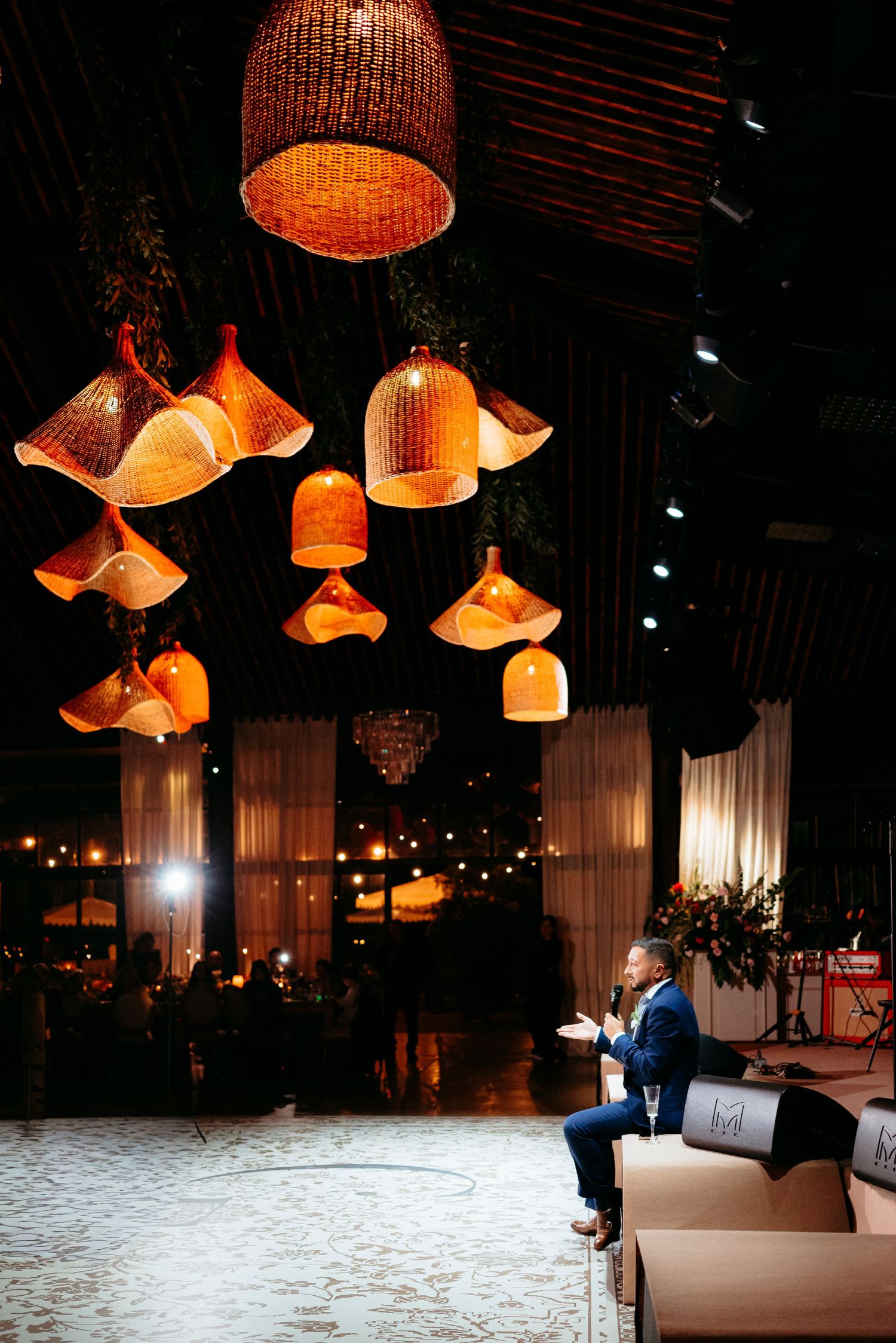 Man in blue suit seated on sofa under ornate hanging wicker pendant lights at elegant evening wedding reception.